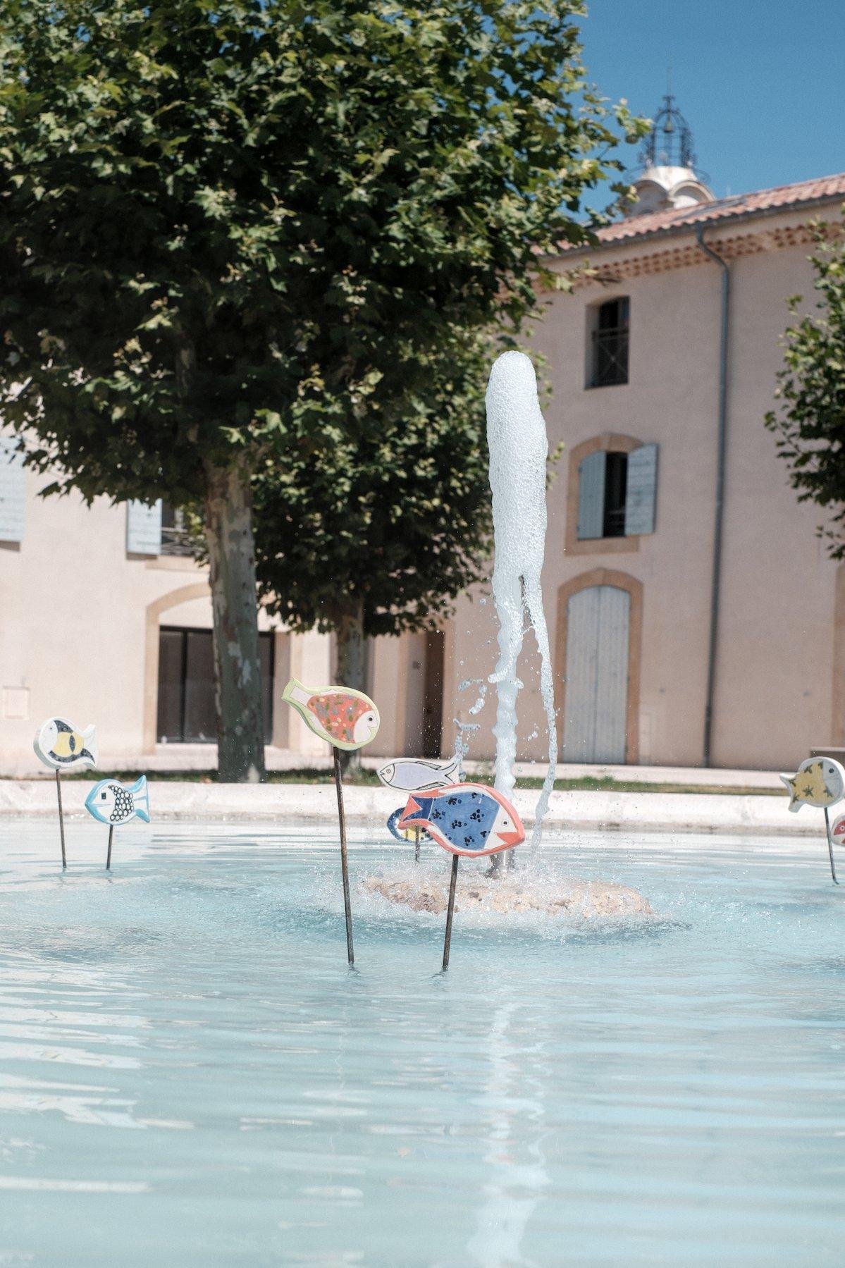 Photo verticale. Une fontaine à l’eau claire avec un petit jet verticale retombant et des petites pancartes en forme de poisson plantées sur des bâtons noirs dans la fontaine les maintenant au dessus de l’eau. Au fond, un gros arbre vert et des bâtiment aux couleurs rosés