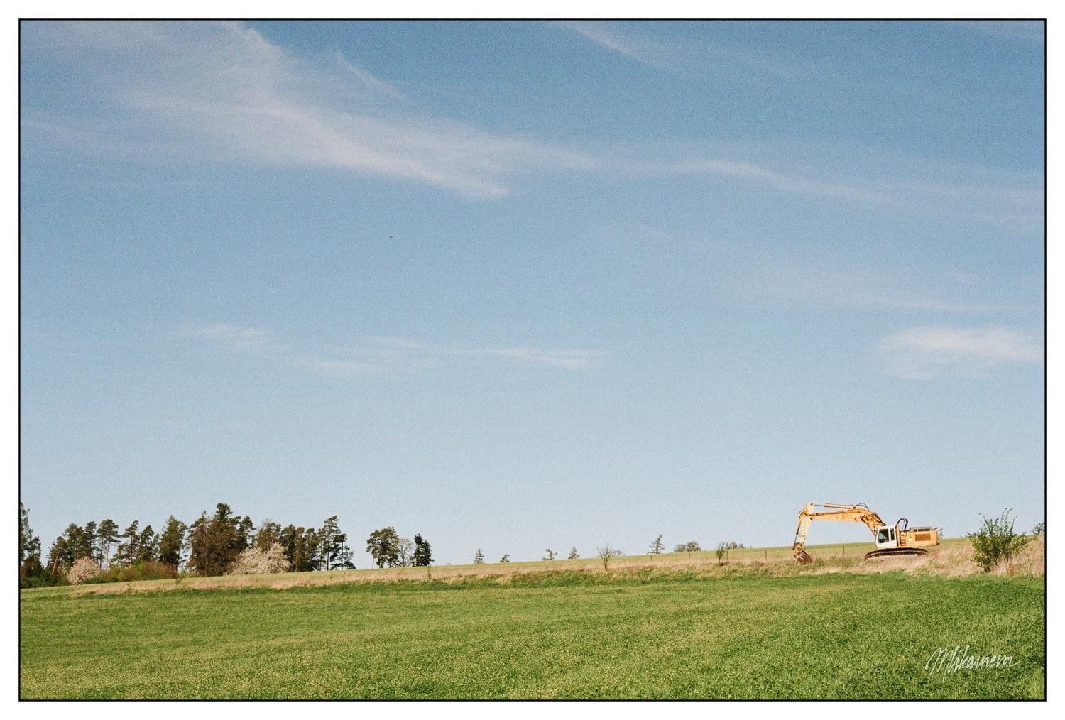 Excavator sits on a grassy field under a clear blue sky with a few clouds. Sparse trees line the horizon in the background on the left side.
