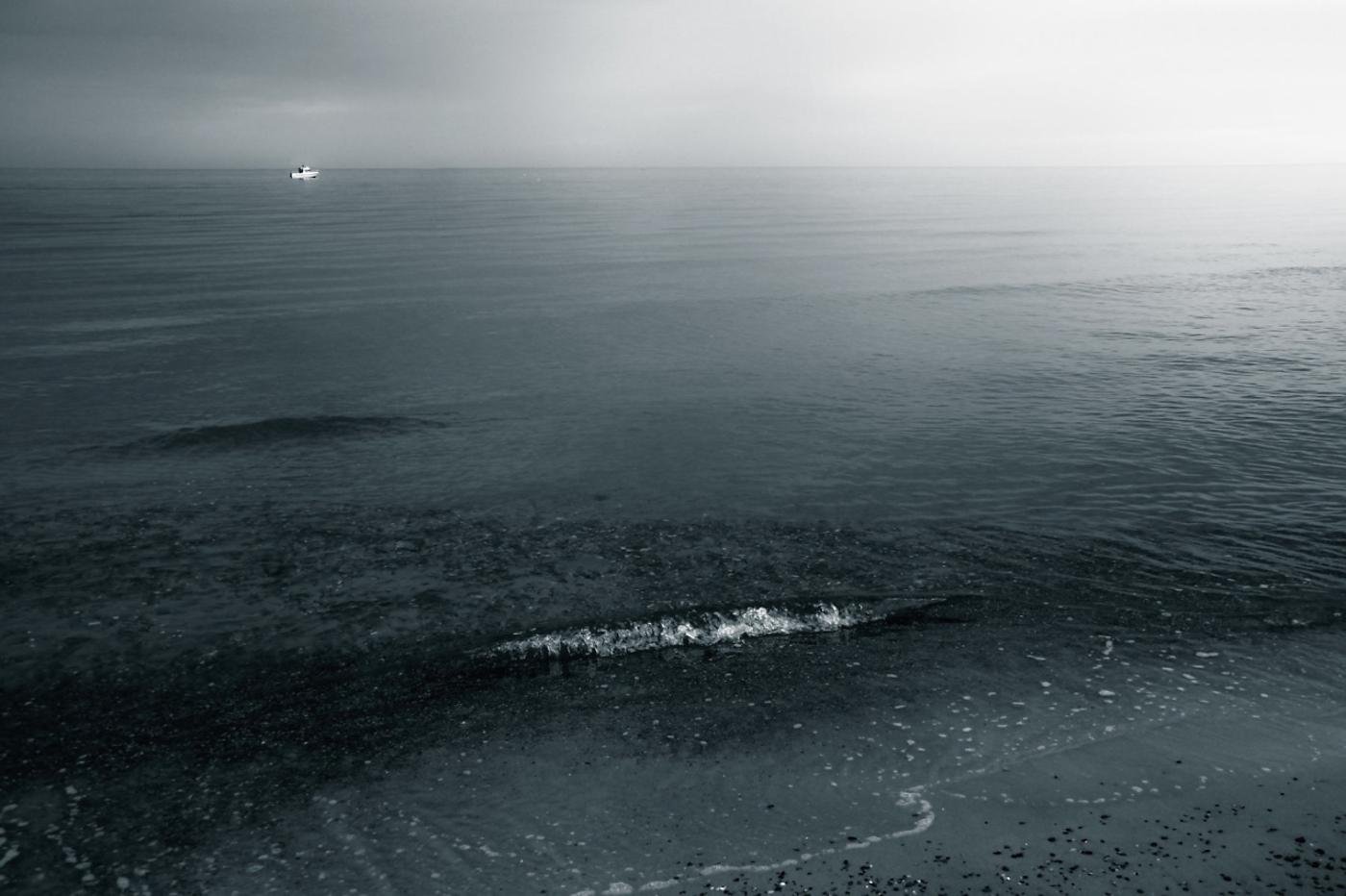 Wide view of a calm ocean under an overcast sky, with a small white boat near the horizon at upper left and a dark band of water with a breaking wave in the foreground.