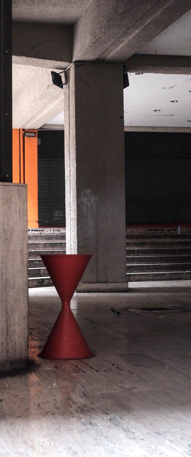 Photo verticale. Dans une galerie avec de la lumière naturelle qui arrive à descendre, un objet orange en forme de sablier. Probablement un cendrier retro