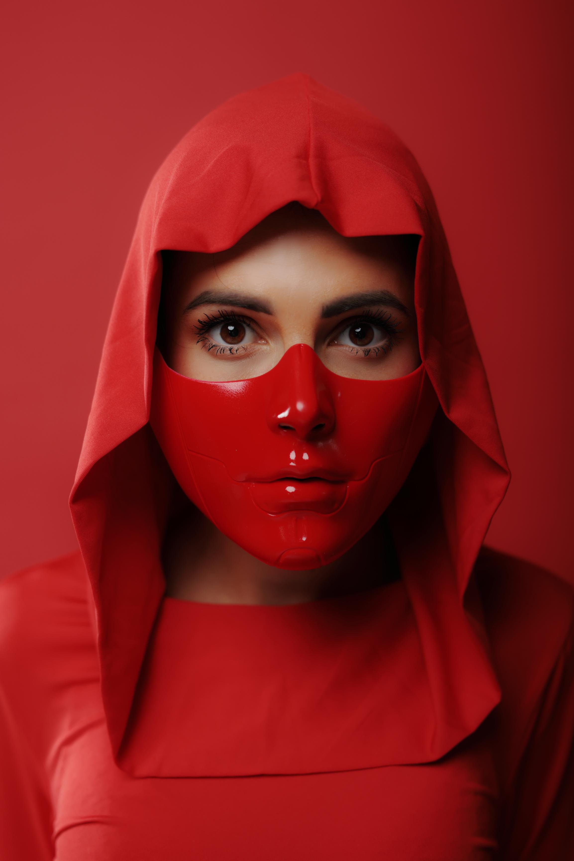 A person wearing a glossy red face mask and a red hooded garment, set against a red background. The eyes are visible and centrally focused. The overall theme is monochromatic in various shades of red.