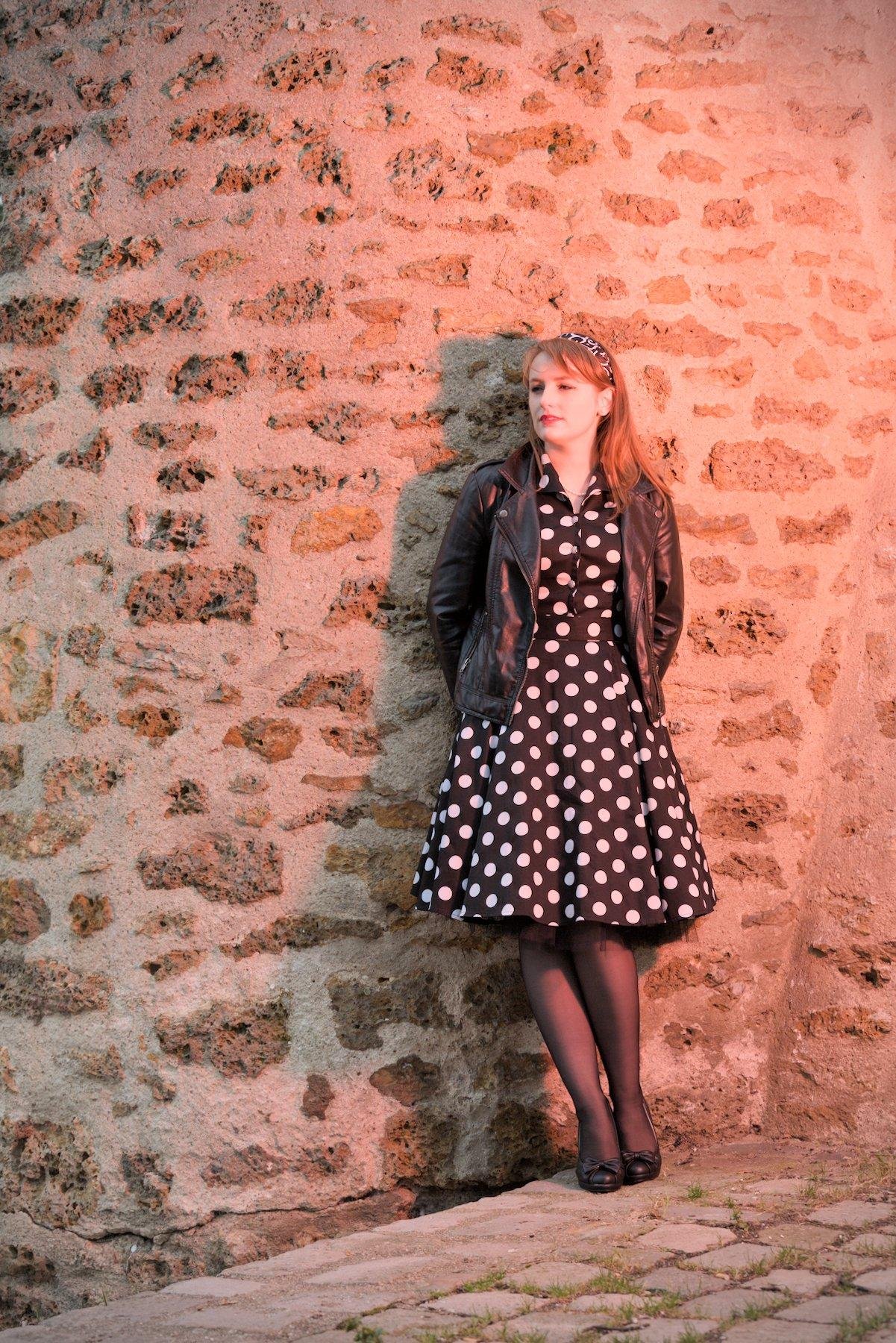 Photo verticale. Une jeune femme aux cheveux long roux, posant devant un mur de vieilles pierres, portant une robe noire à pois blanc, des collants noirs, des chaussures noires avec un petit nœud, un blouson en cuir noir et un serre tête noir avec des motifs blancs. Ses mains son derrières elle. Elle regarde au loin vers sa droite, le sol est très penché et pavé. La scène est dans les teintes rouges/rose