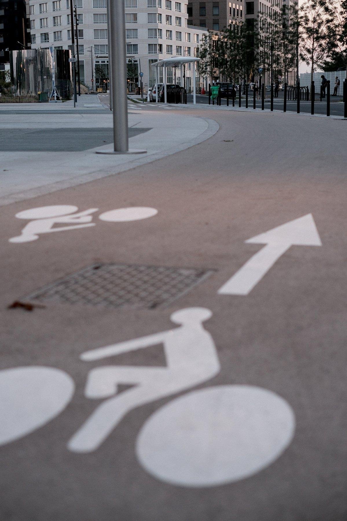 Photo verticale. à ras du sol d’une piste cyclable avec le symbole de cycliste et une flèche, on voit la piste s’étirer en serpentant vers des immeubles blanc éclairés par le soleil couchant