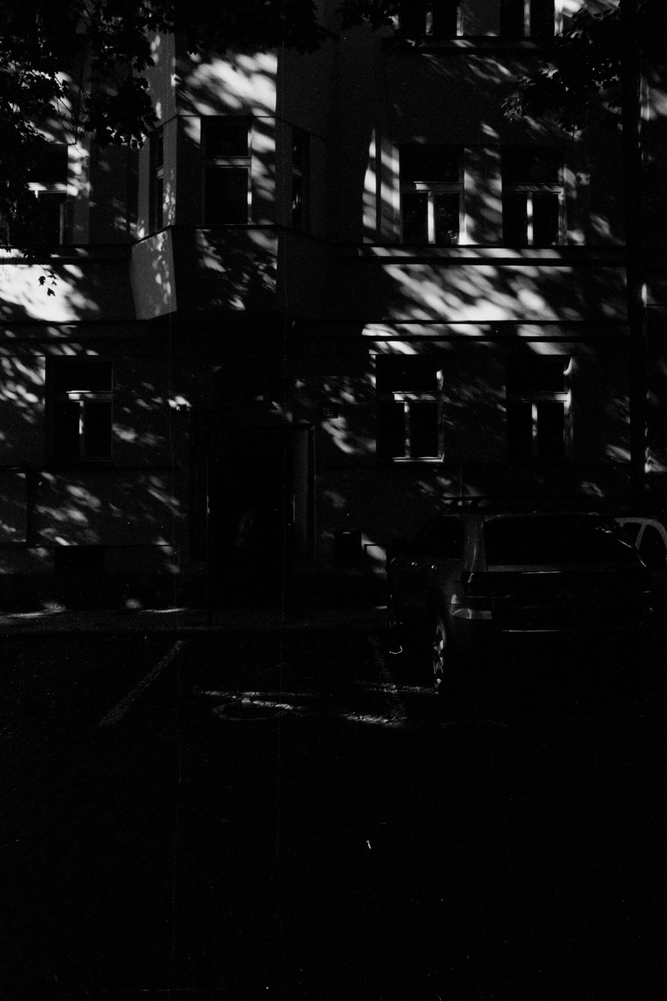 Black-and-white photo of a multi-story apartment building facade with several rectangular windows; dappled shadows from tree branches fall across the walls, and a parked car is partially visible at the lower right.