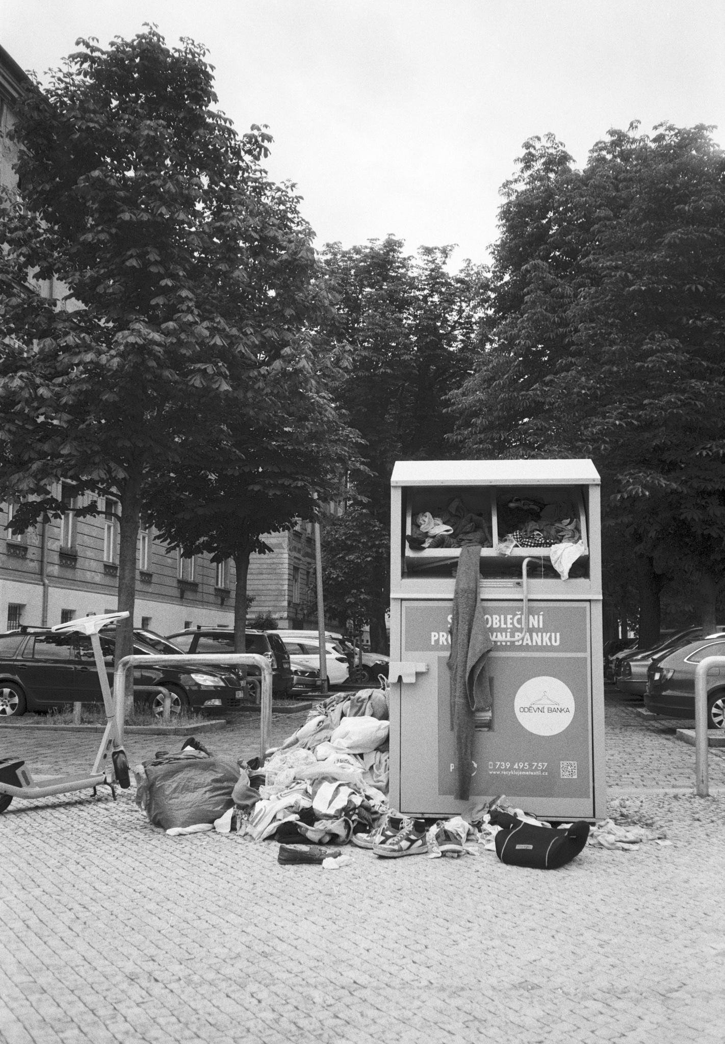Overflowing clothing donation bin on a cobblestone sidewalk, with piles of discarded clothes, shoes, and bags scattered around the base; parked cars and leafy trees line the street beside an apartment building in the background.