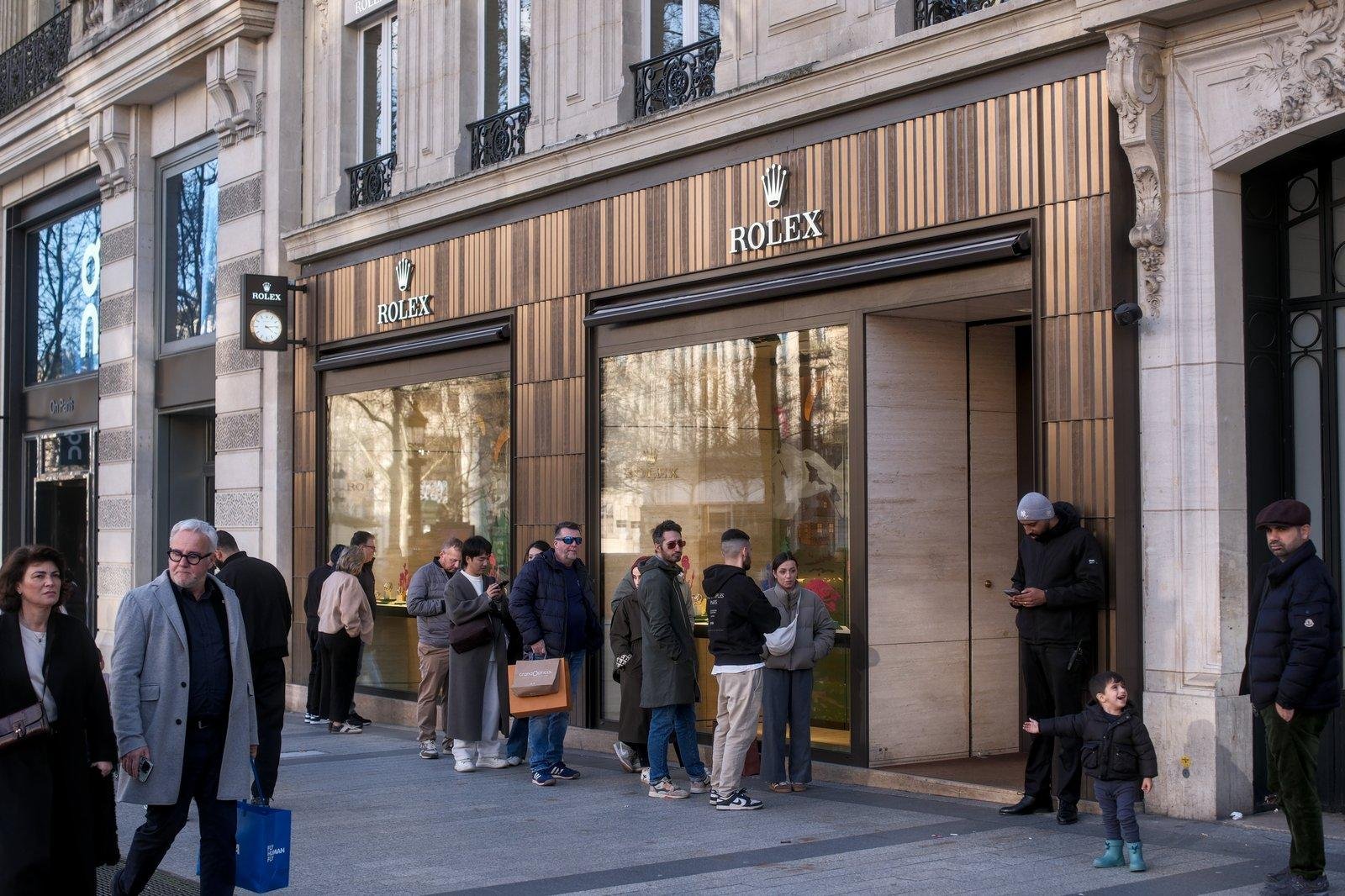 Des gens faisant la queue pour entrer dans un magasin Rolex. Sans aucun rapport avec eux, à droite de l’entrée, un enfant à la mine réjouie montre quelque chose de la main à son père qui ne regarde pas
