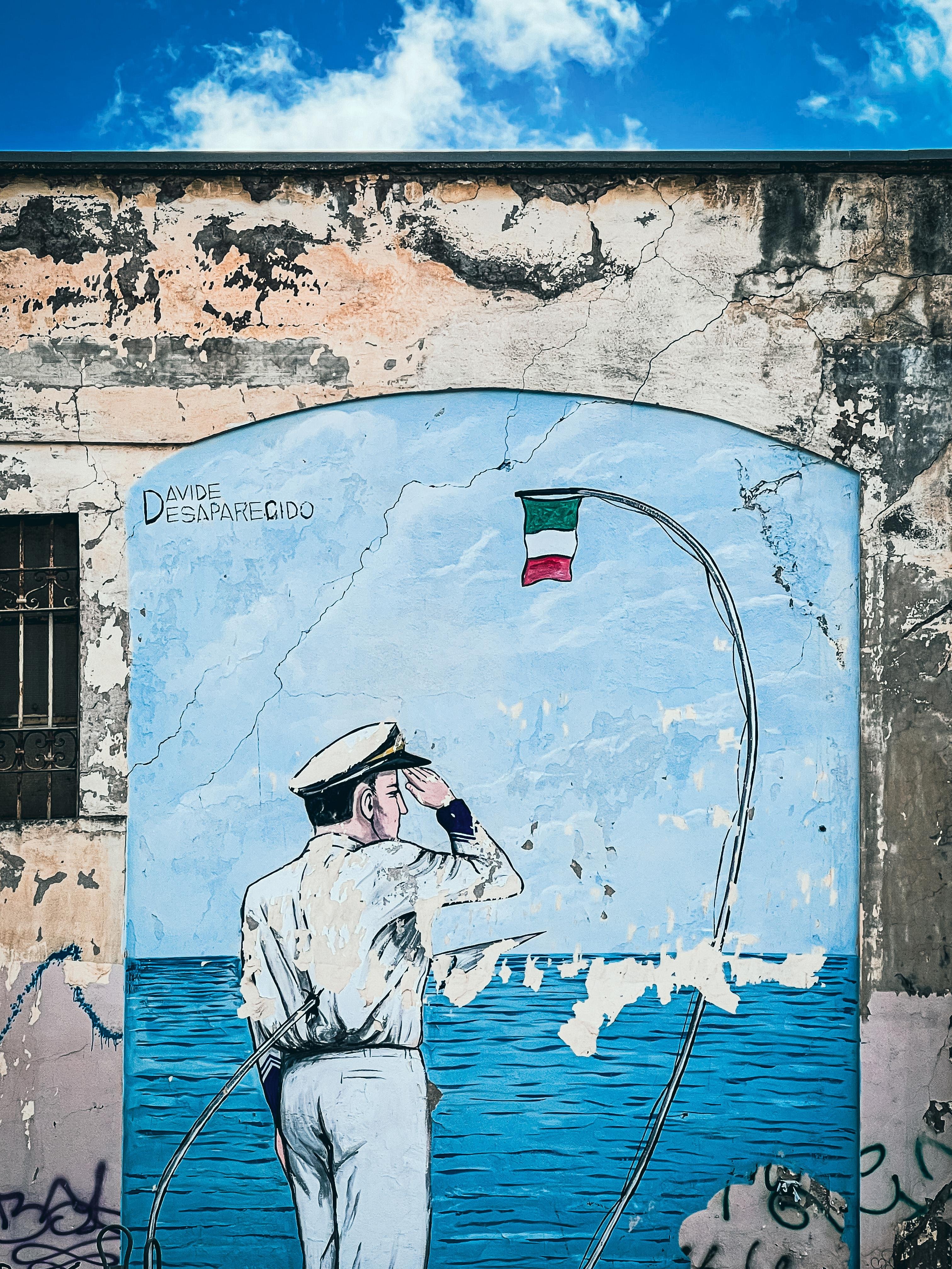 Murale su un muro logoro raffigurante un marinaio in uniforme che sta in piedi e saluta verso il mare. Sopra, c'è una piccola bandiera italiana su un'asta piegata. Il muro intorno al murale sembra scheggiato e danneggiato. Le parole "Davide Desaparecido" sono scritte sul murale.