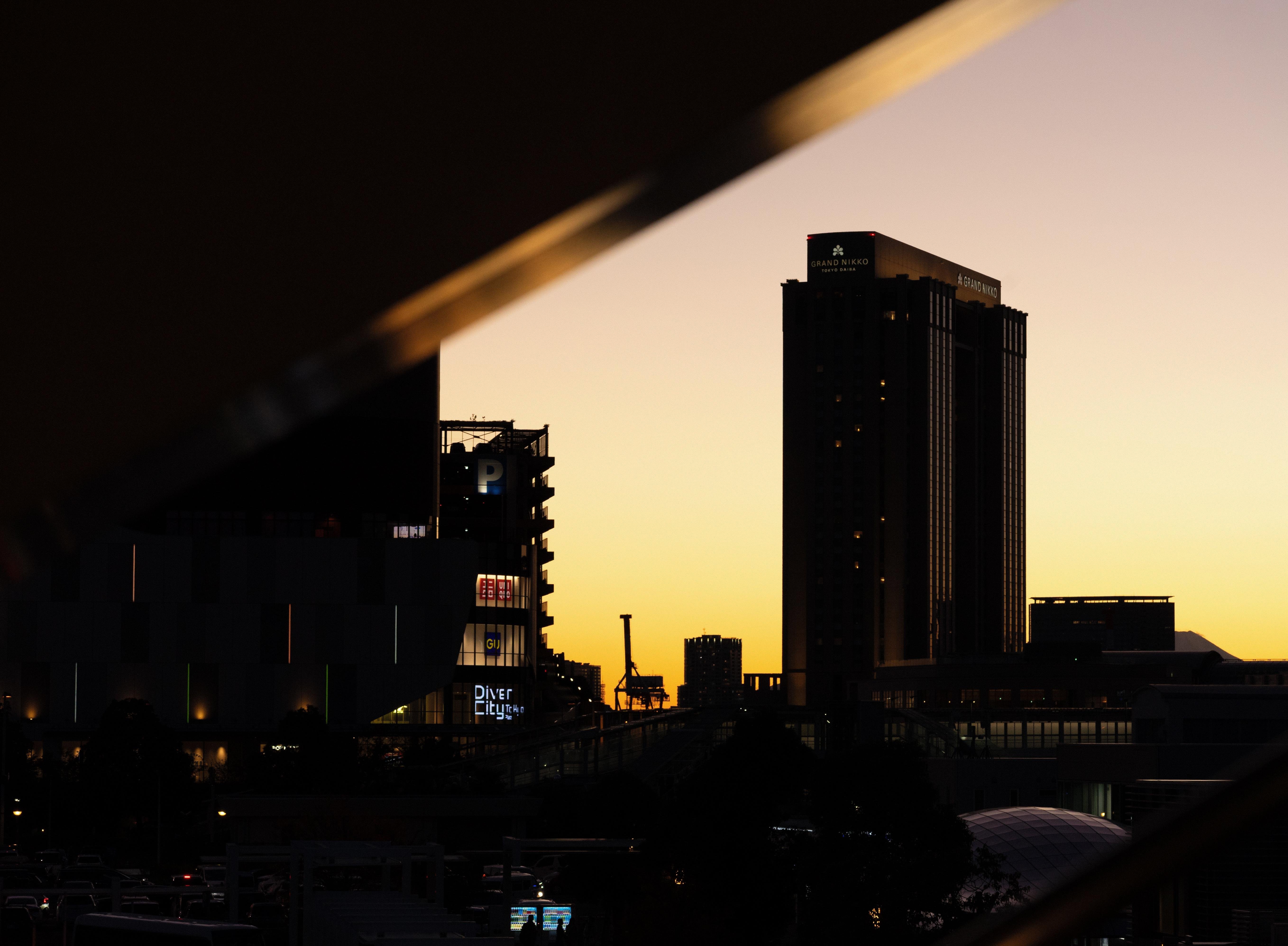 Silhouetted city skyline during sunset, featuring tall buildings with visible signs. The sky transitions from deep orange to purple hues. Geometric structure frames the upper corners of the image.