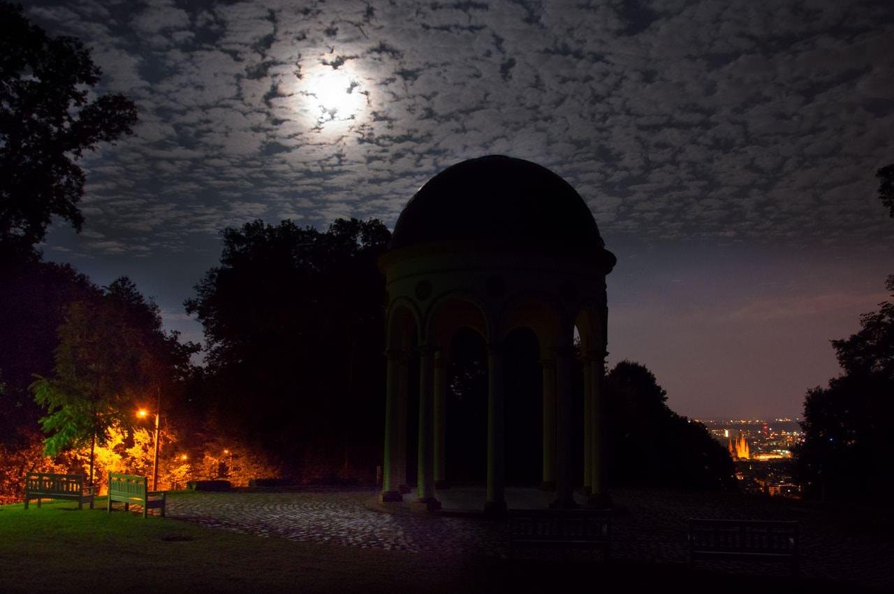 Moonlit night scene with a domed pavilion in silhouette at center, trees framing the park, a lit streetlamp and benches on the left, cobblestone path in the foreground, and distant city lights on the right under a cloudy sky.
