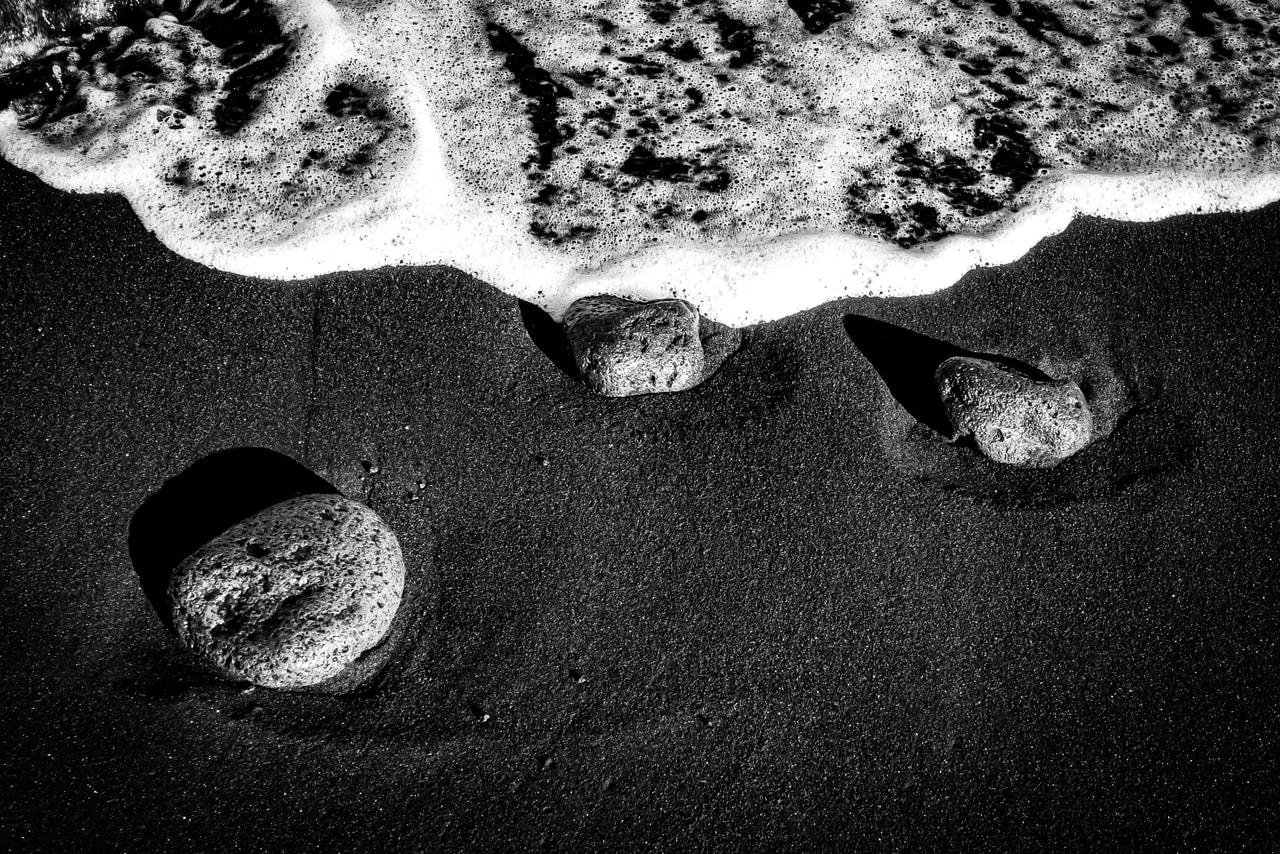Black-and-white close-up of foamy surf along the top edge meeting dark wet sand, with three rounded stones partly embedded in the sand casting long shadows toward the right.