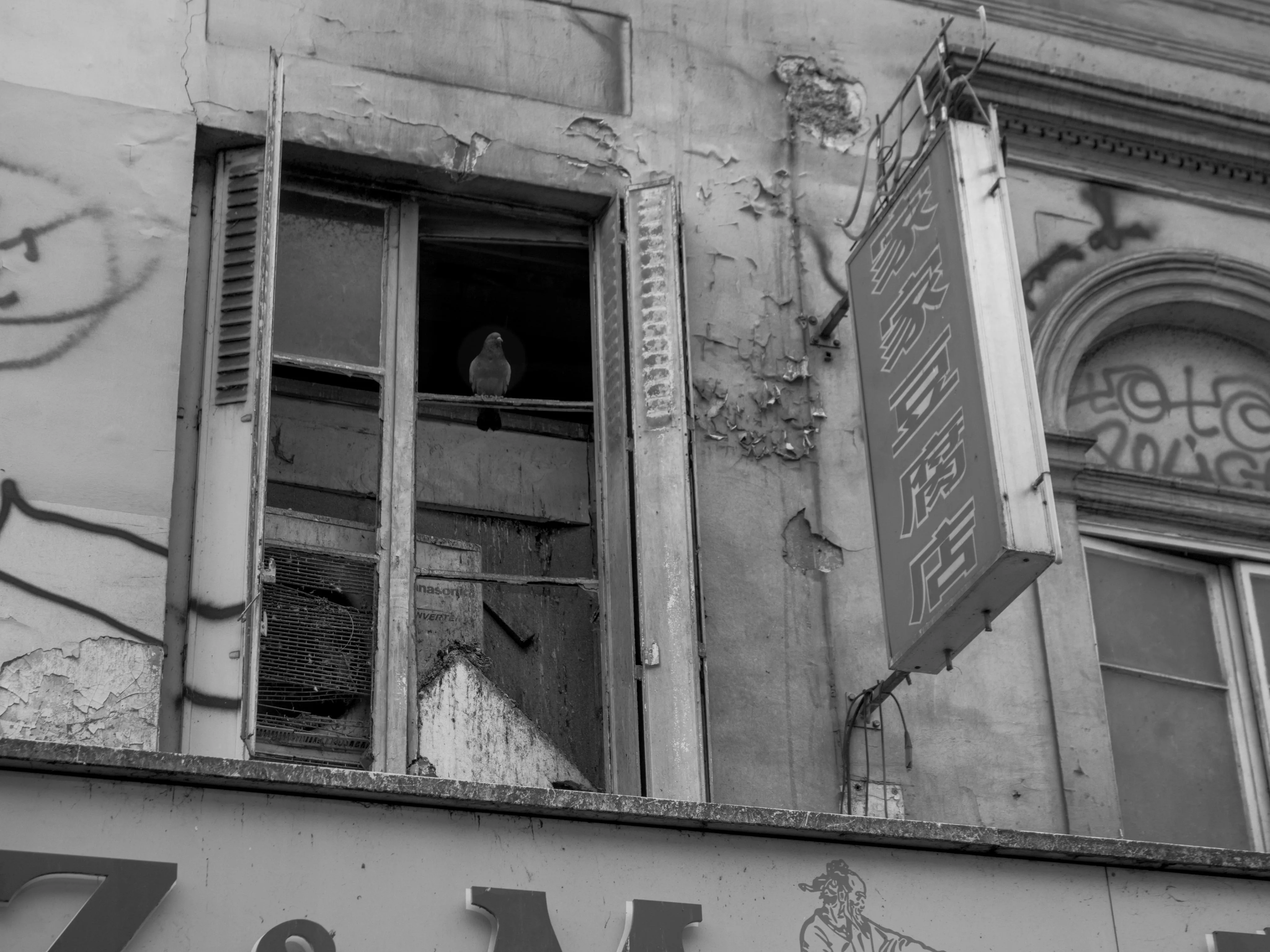 Photograph of a pigeon sitting on the bar of a broken window of a seemingly abandoned building. The photograph is taken from a lower level than the window, at a high angle. An old air conditioner-like machine sits inside the window with a lot of bird dropping running down its surface.  Beside the window, a street sign with Chinese characters indicates a restaurant or store.