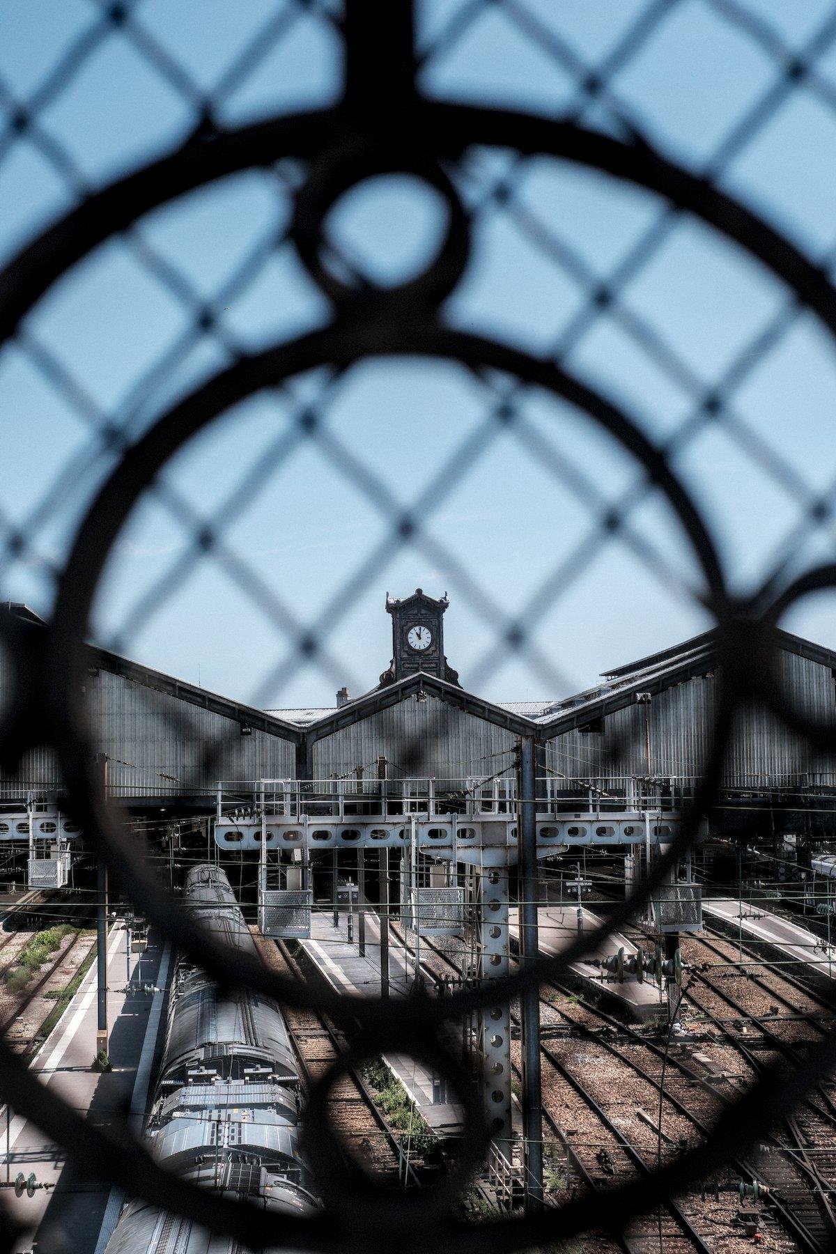 Photo verticale. À travers un grillage derrière une barrière en fer forgé, l’horloge d’une gare au sommet des hangars abritant les quais