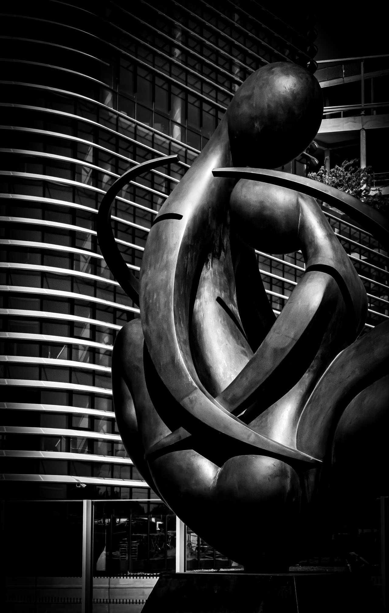 Abstract bronze sculpture with smooth, intertwined forms set against a modern building facade featuring vertical and horizontal lines. The contrast between the sculpture's curves and the building's structured grid creates a dynamic visual. The image is in black and white, highlighting textures and shadows.