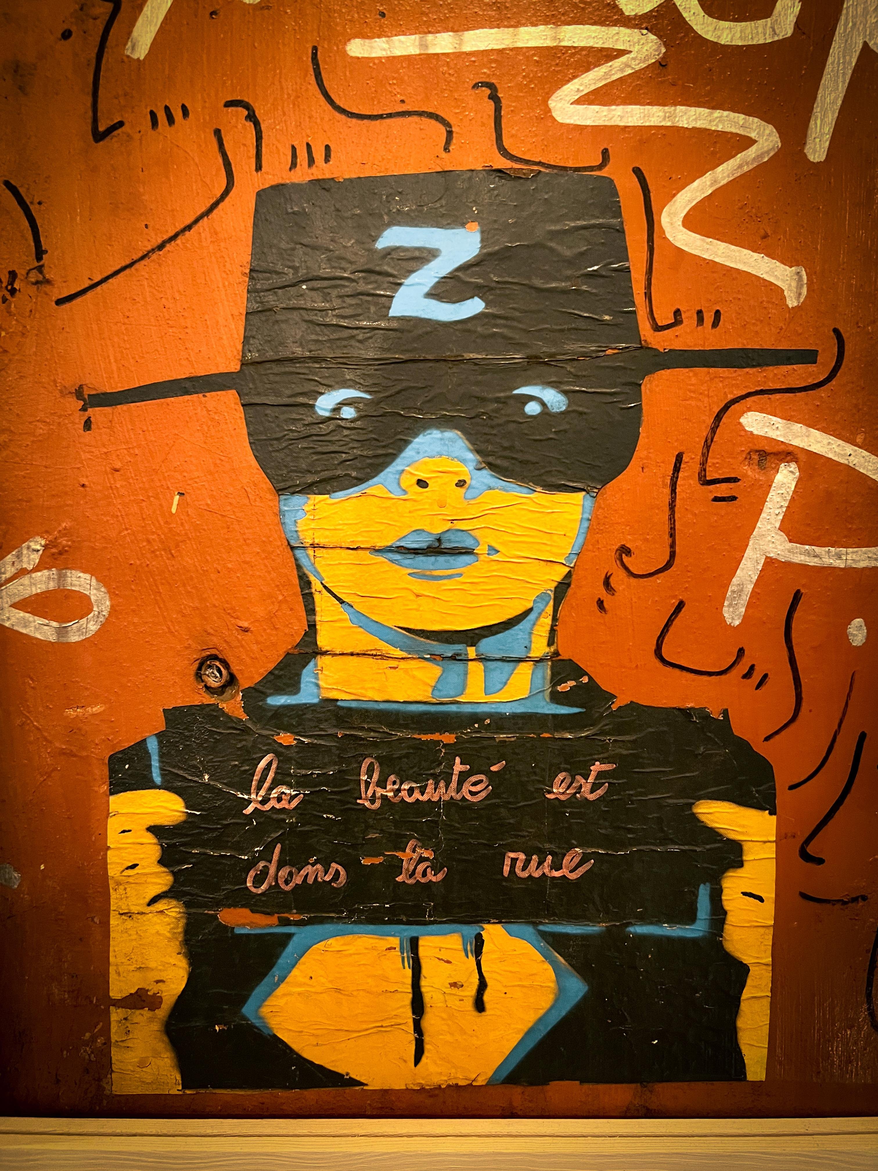 Elemento di arte di strada raffigurante una figura stilizzata che indossa un cappello e una maschera con la lettera “Z”. L’opera d’arte include colori vivaci come blu, giallo e nero su uno sfondo arancione. La figura tiene un cartello con la frase francese “la beauté est dans la rue,” che si traduce in “la bellezza è nella strada.” L’area circostante ha linee e forme astratte.