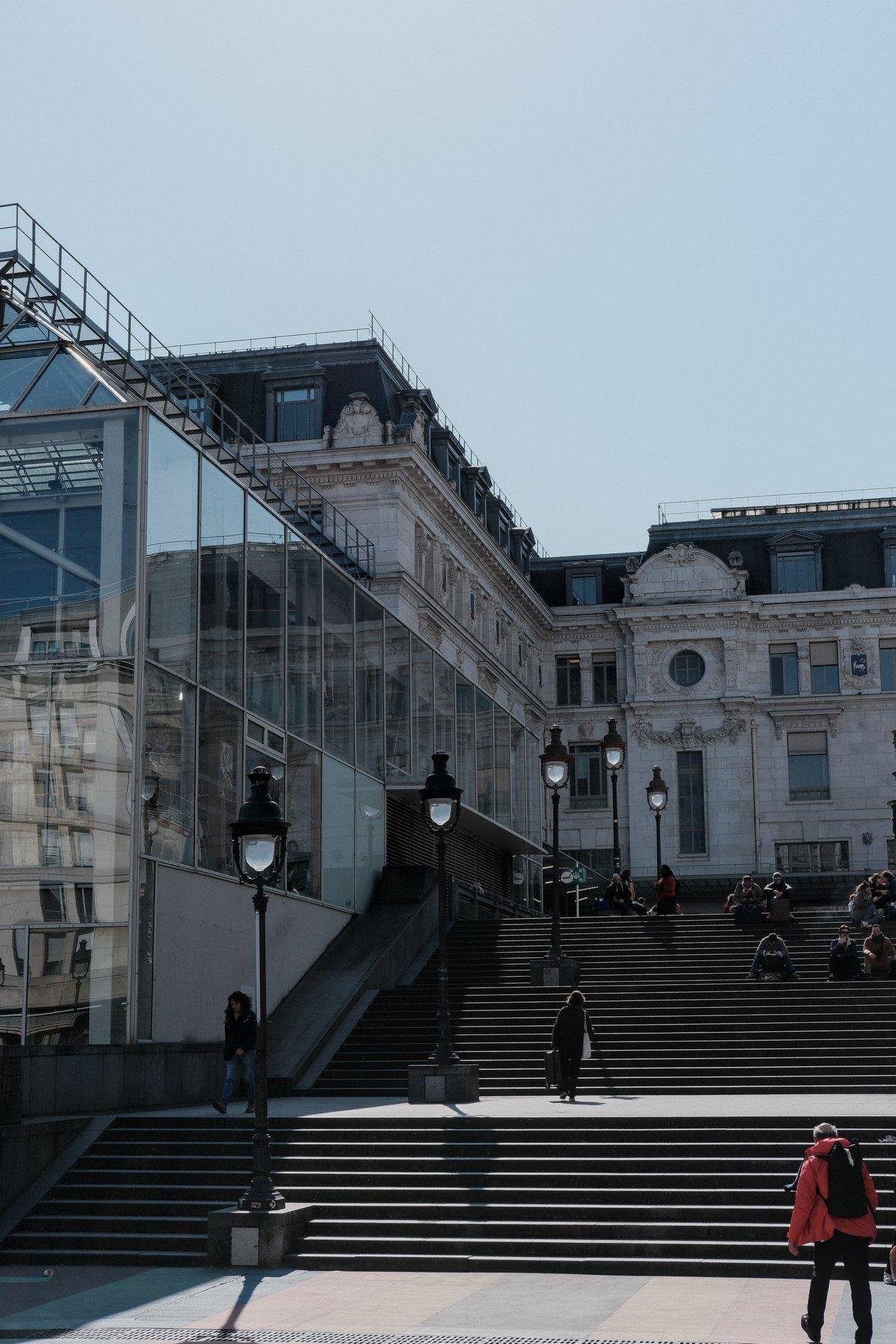 Photo verticale. Un très large escalier en avec 2 paliers et des lampadaires réguliers, à droite d’un bâtiment élégant une partie en architecture classique et l’autre en moderne en verre. Des gens l’empruntent