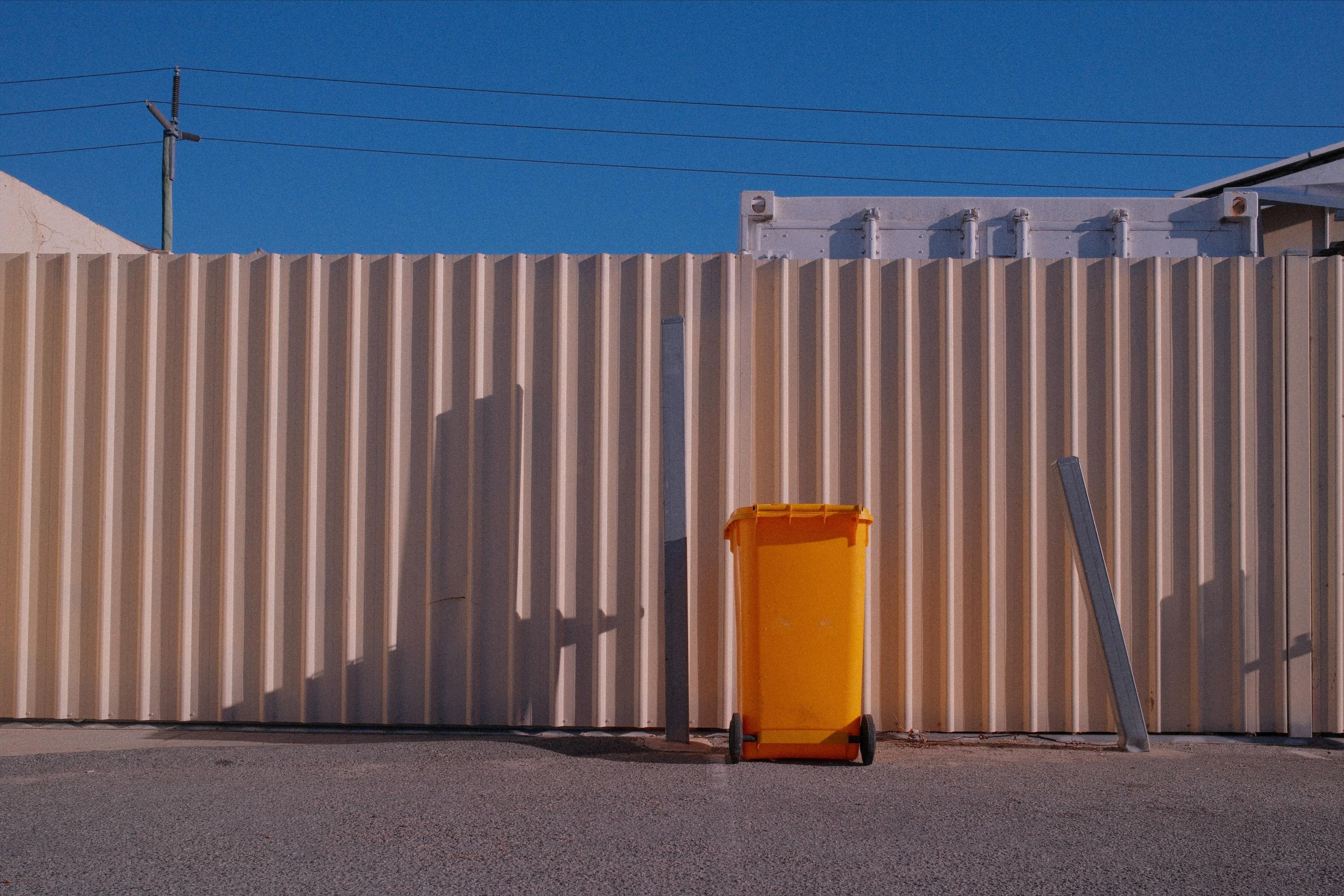 Yellow wheelie bin on asphalt in front of a beige corrugated metal fence, with two leaning metal posts nearby and utility wires against a clear blue sky because Perth.