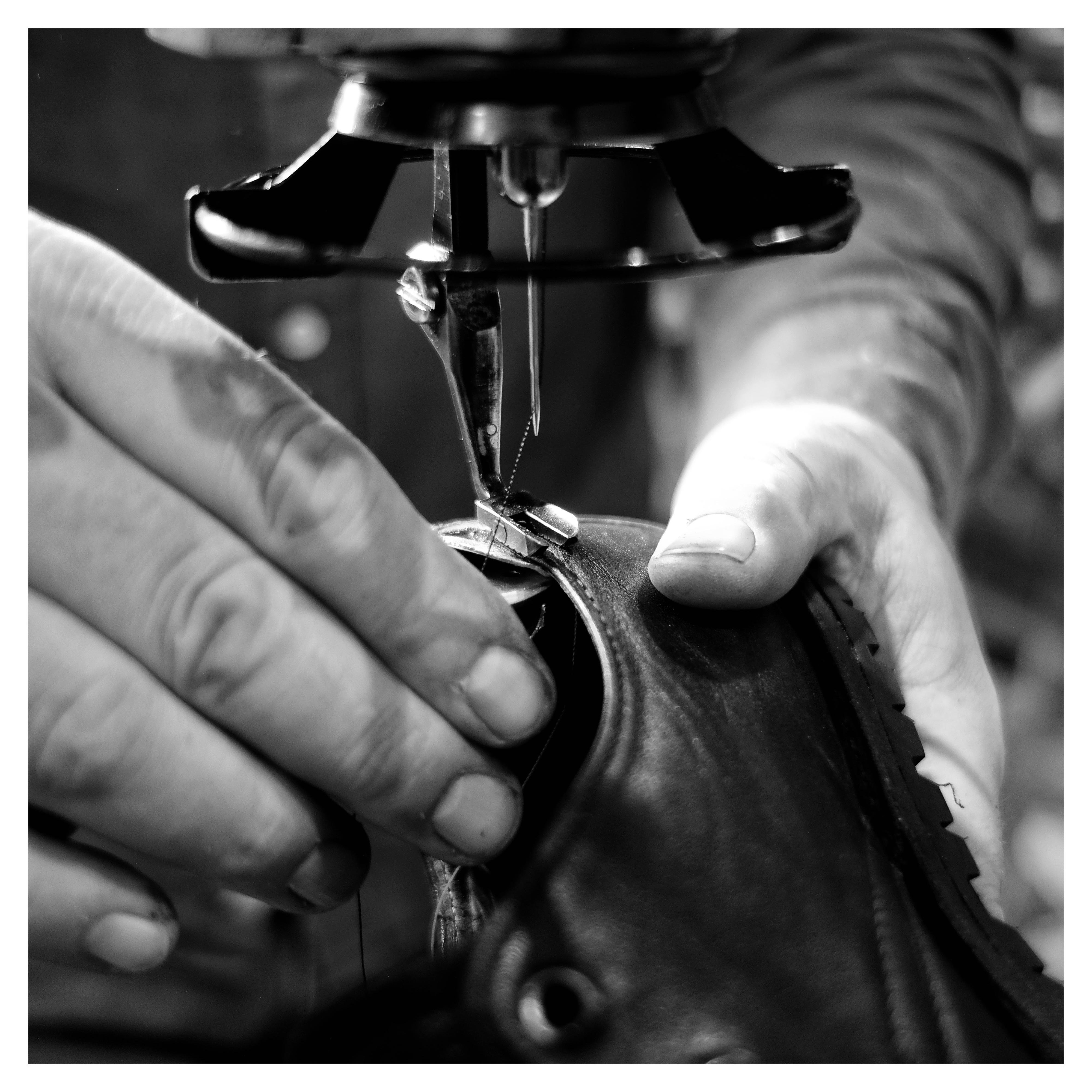 Close-up black-and-white photo of hands guiding a leather shoe under an industrial sewing machine needle, stitching along the shoe’s upper near the sole.