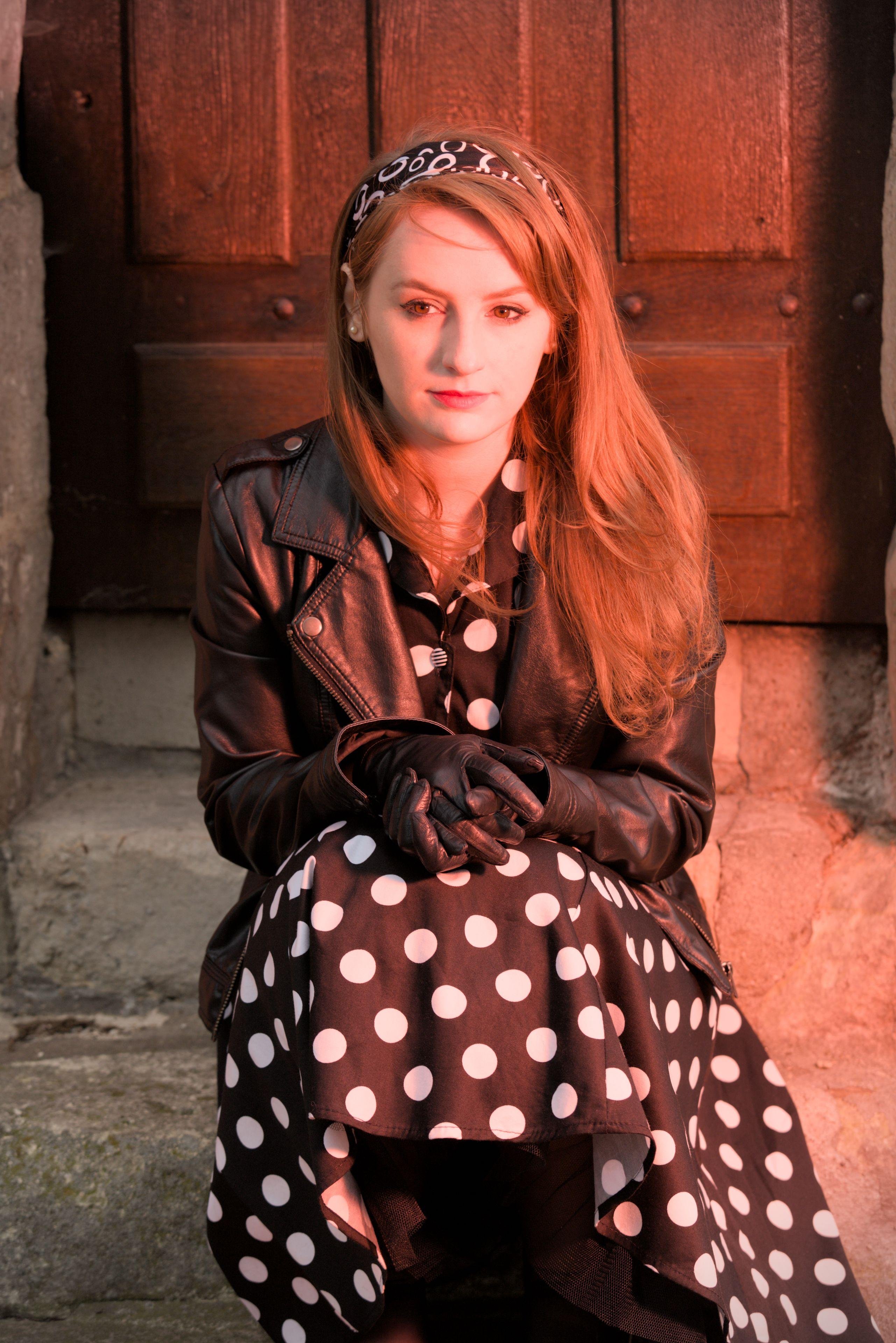 Une fille assise sur les marches d’un petit escalier en pierre, devant une porte, la tête de face mais les yeux regardant vers sa droite, les mains posées sur ses genoux, une expression rêveuse/mélancolique/songeuse,