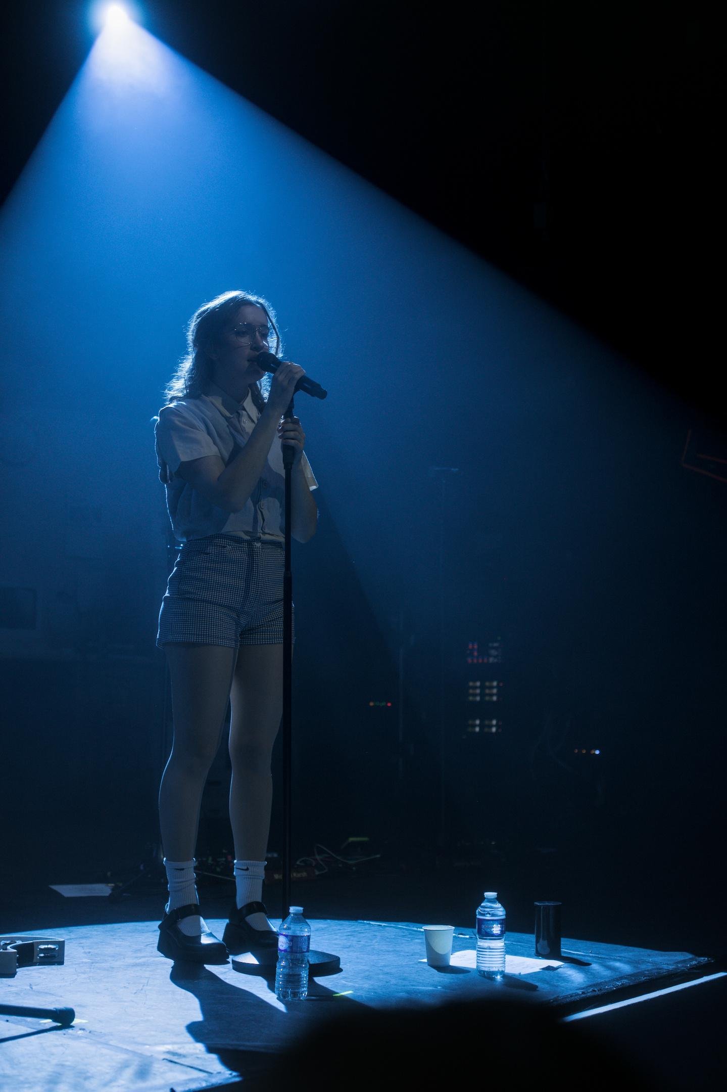Photo verticale. Gracie Lawrence sur scène, debout devant son micro, un cône de lumière bleu l’encadre