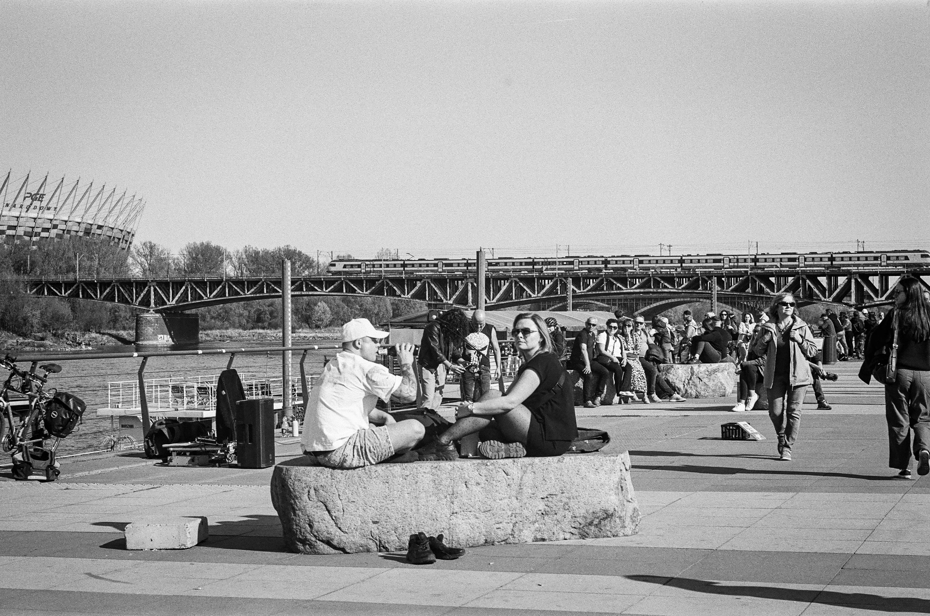 B&W photo of a couple sitting on a rock at Vistula boulevards in Warsaw. Behind them, there are street performers singing and a train passing on a railway bridge in the background