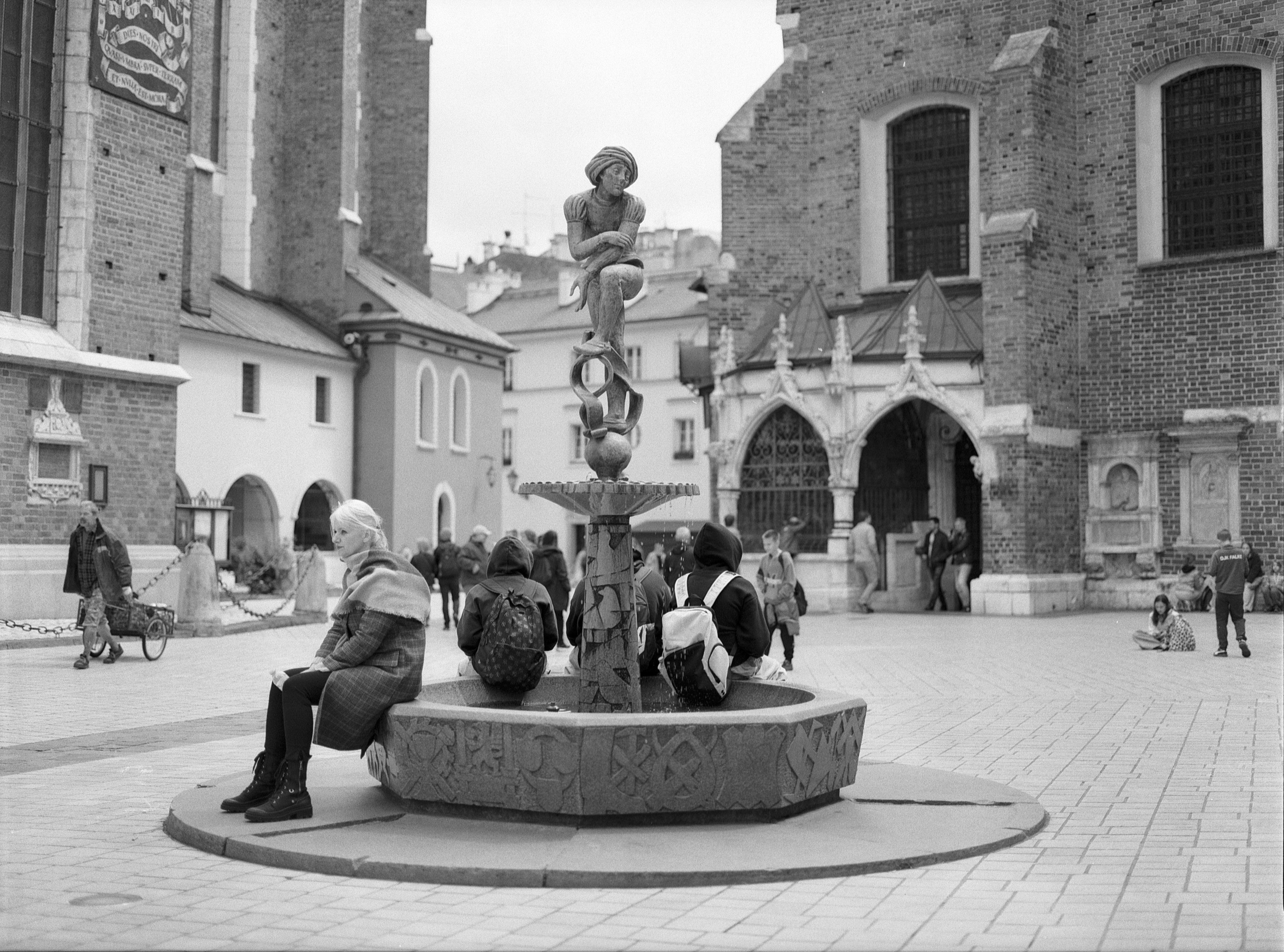 Sculpture fountain in a public square with people sitting around its base. The fountain features a seated figure atop an intricately designed column. A large brick building with arched windows in the background, along with smaller surrounding structures. People are scattered throughout the square, including individuals near a building entrance and a child playing on the ground. The scene is captured in black and white, highlighting the architectural details and cobblestone pavement.