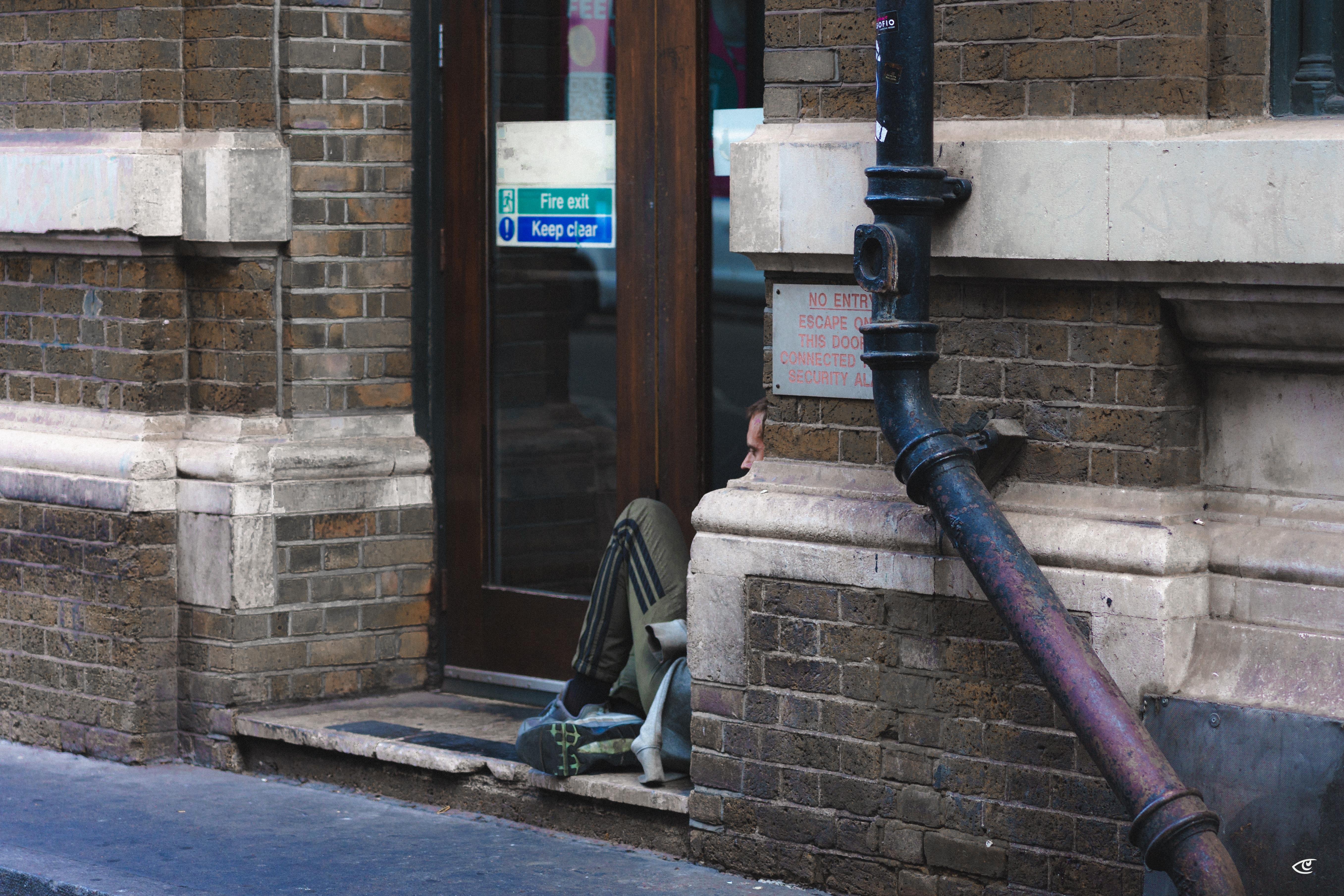 Person sitting on the ground beside a doorway in a brick building, partially hidden behind a stone corner, with a bag and clothing on the pavement; glass door behind has a “Fire exit keep clear” sign and a large black drainpipe runs along the wall.