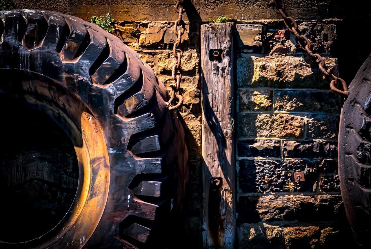 The image shows large, rugged tires hanging on a wall. The wall is made of stone, and there is wooden plank and metal chain attached to it. The tires appear to be used as bumpers, possibly for a dock or similar setting.