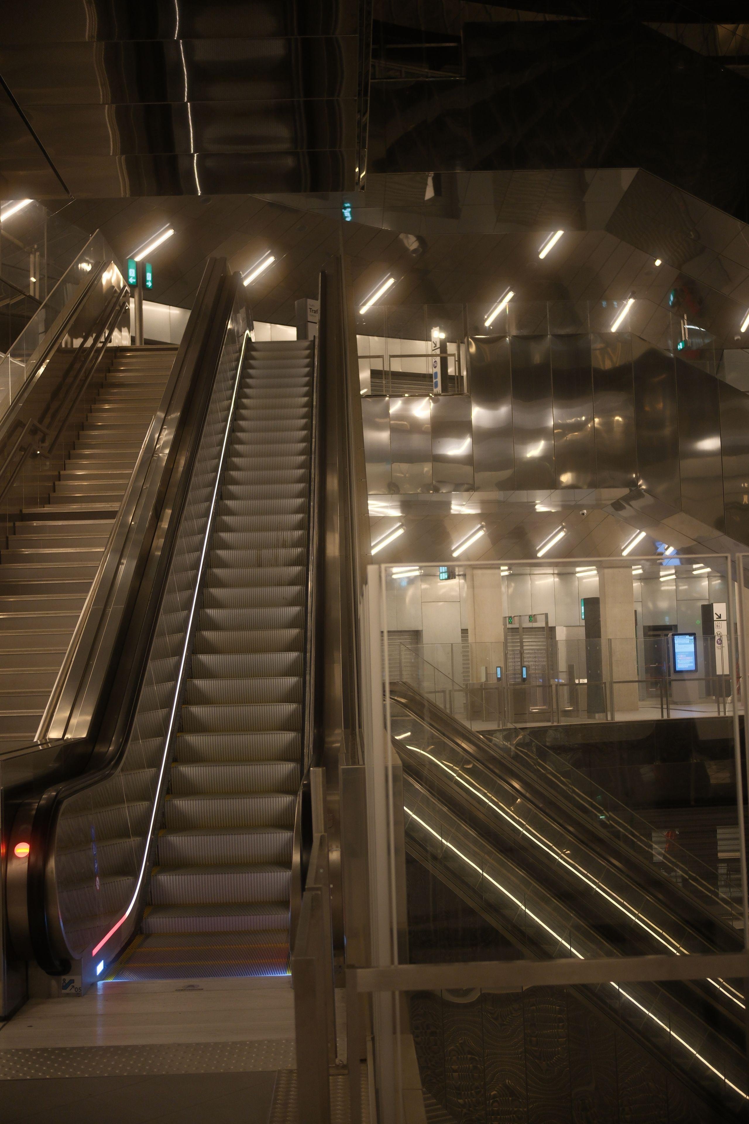 A pic of mechanical stairs, one faced from the bottom of it, and behind it, another one lateral. The place looks huge and a metallic light orange hue is all around because of the metallic covers on a lot of walls
