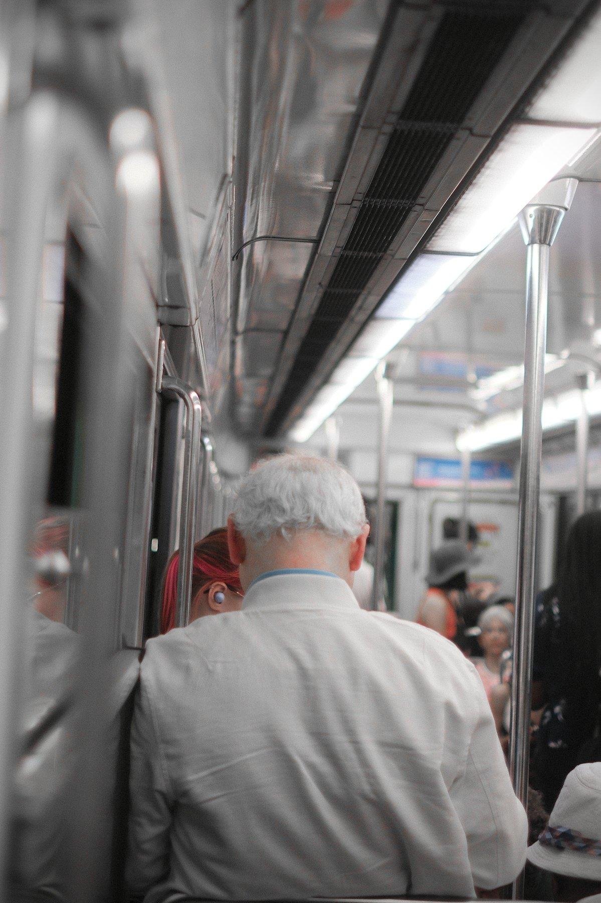 Photo verticale. Dans une rame de métro, un homme de dos avec des cheveux blanc, blouson blanc, tête un peu baissé (sûrement en train de consulter son tél) et dans l’espace entre la gauche de sa tête, son épaule et les portes du wagon, un bout de tête d’une jeune femme brune aux mèches rose fluo