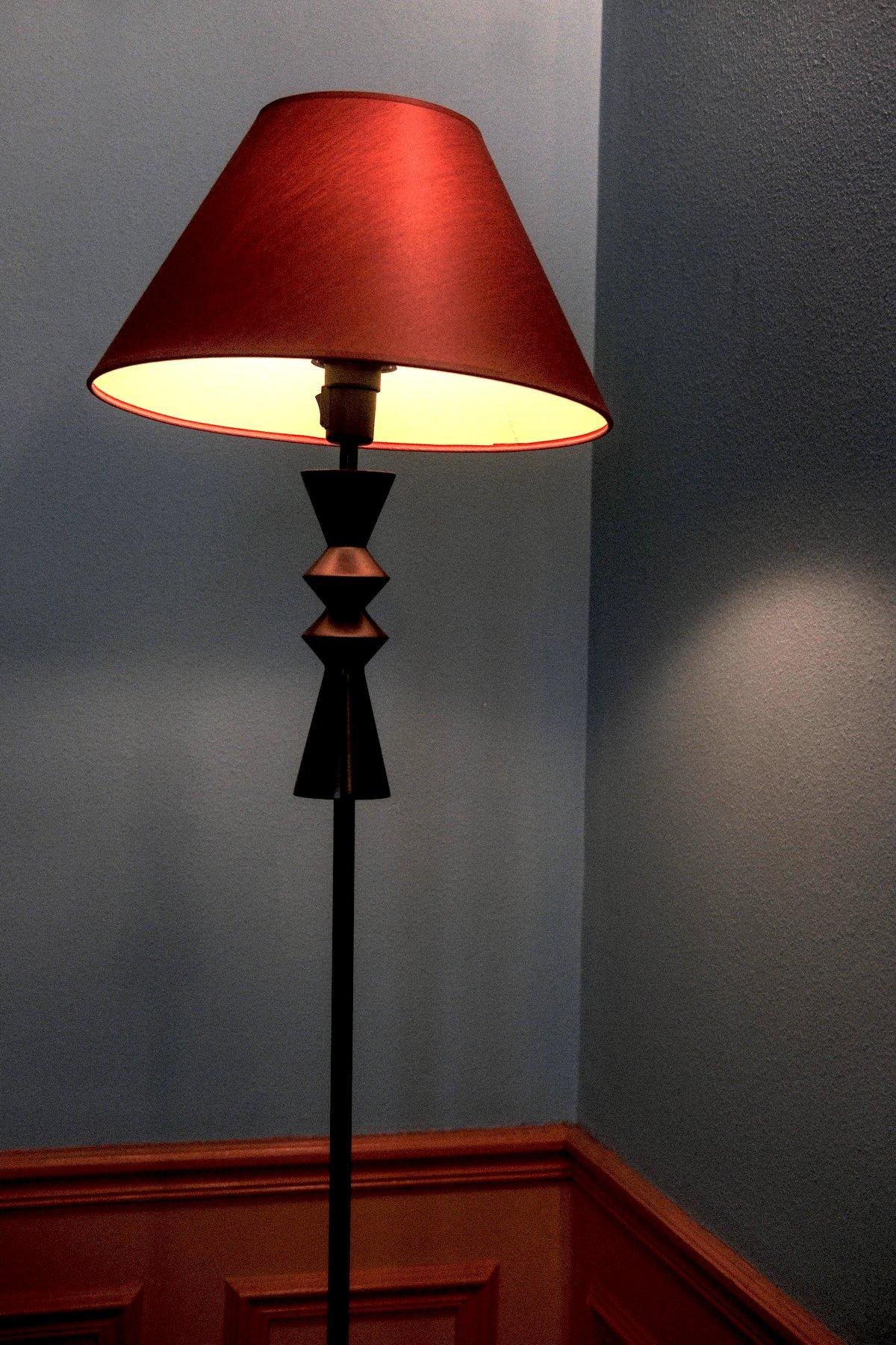 Photo verticale. Un lampadaire allumé de avec un abat jour orange un peu penché, dans l’angle d’une pièce au mur bleu dont le bas est avec des moulures oranges
