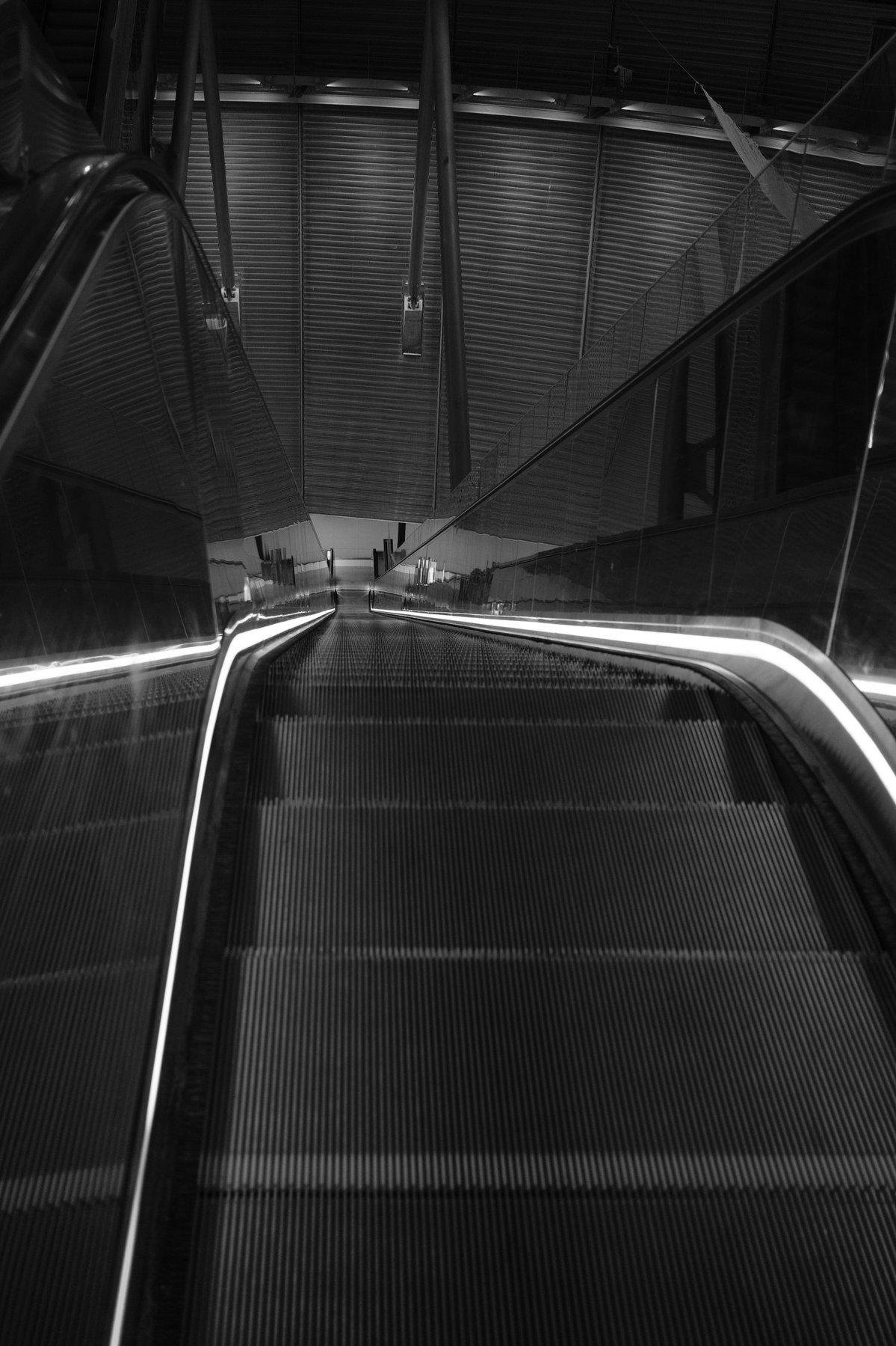 Photo verticale en noir et blanc. Un escalator qui descend, éclairé des deux côtés par une bande de LED. En bas on voit un mur circulaire