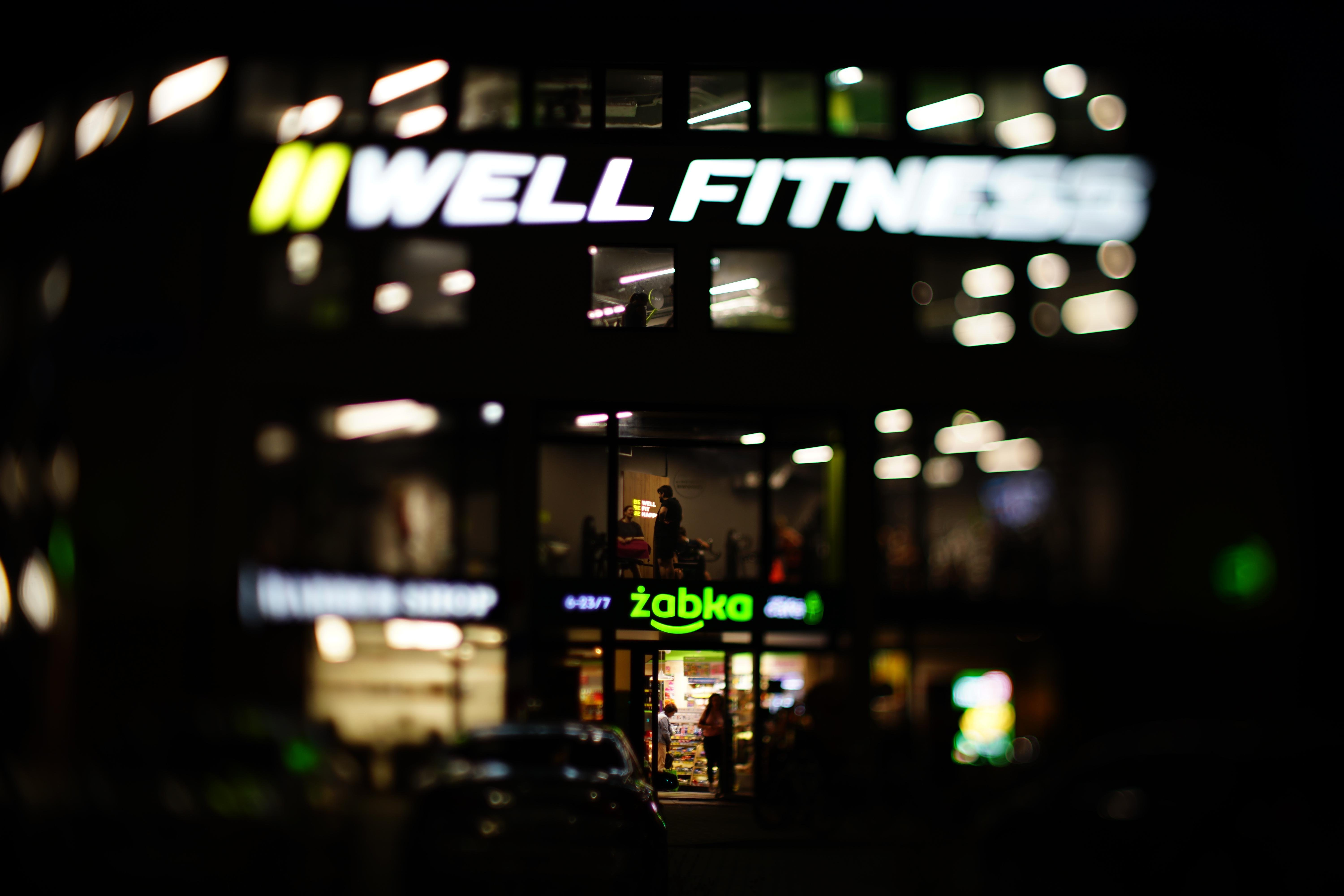 Illuminated building at night with a large "Well Fitness" sign on the upper level and a "Zabka" convenience store on the ground floor. The interior lights create a warm glow, silhouetting people inside. The image is softly focused, emphasizing the vibrant signage.