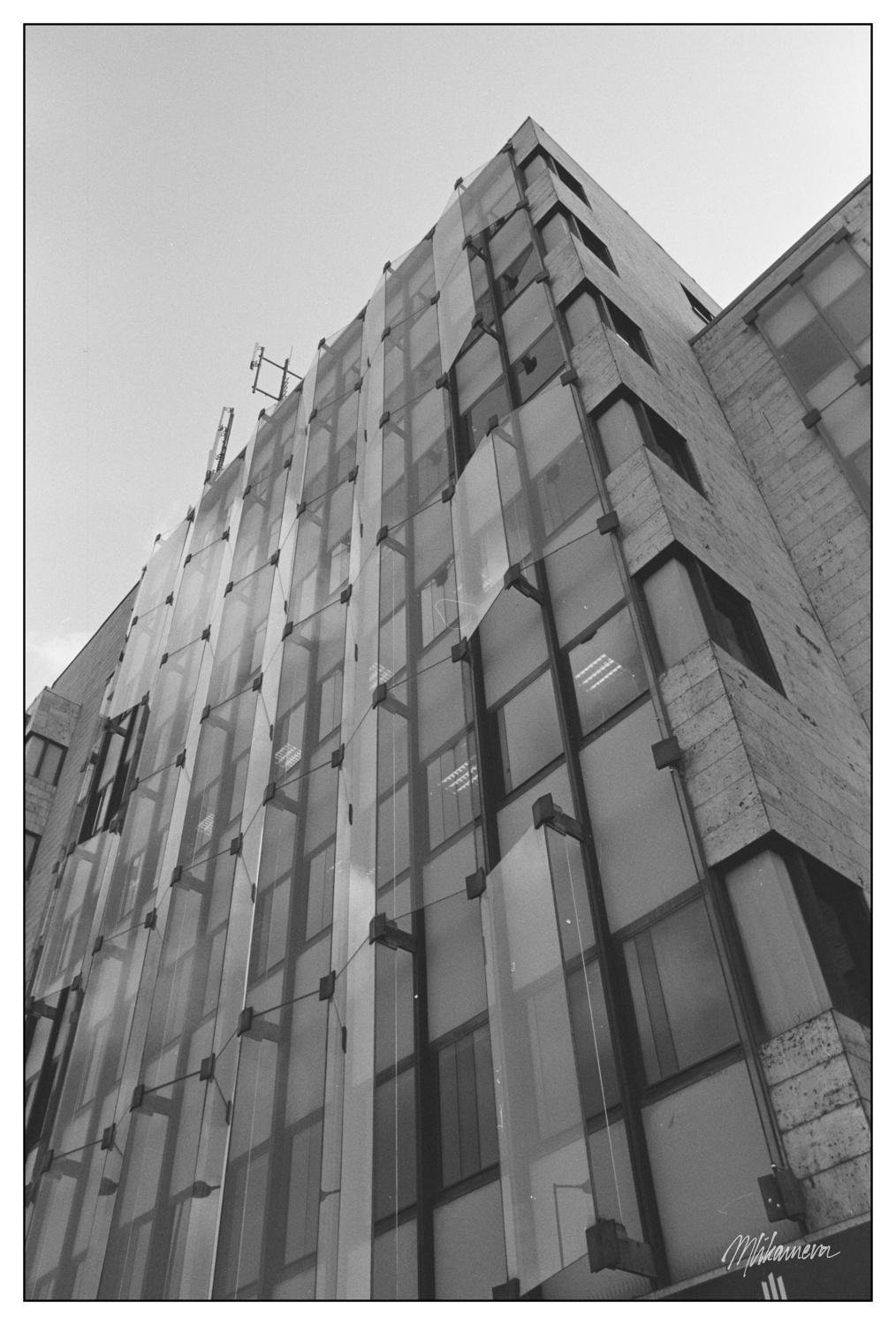 Black and white image of a modern building with a geometric façade composed of large, overlapping glass panels and a concrete structure. The perspective is upward, capturing the building’s vertical lines and architectural details against a clear sky. The glass panels are secured with visible metal brackets.