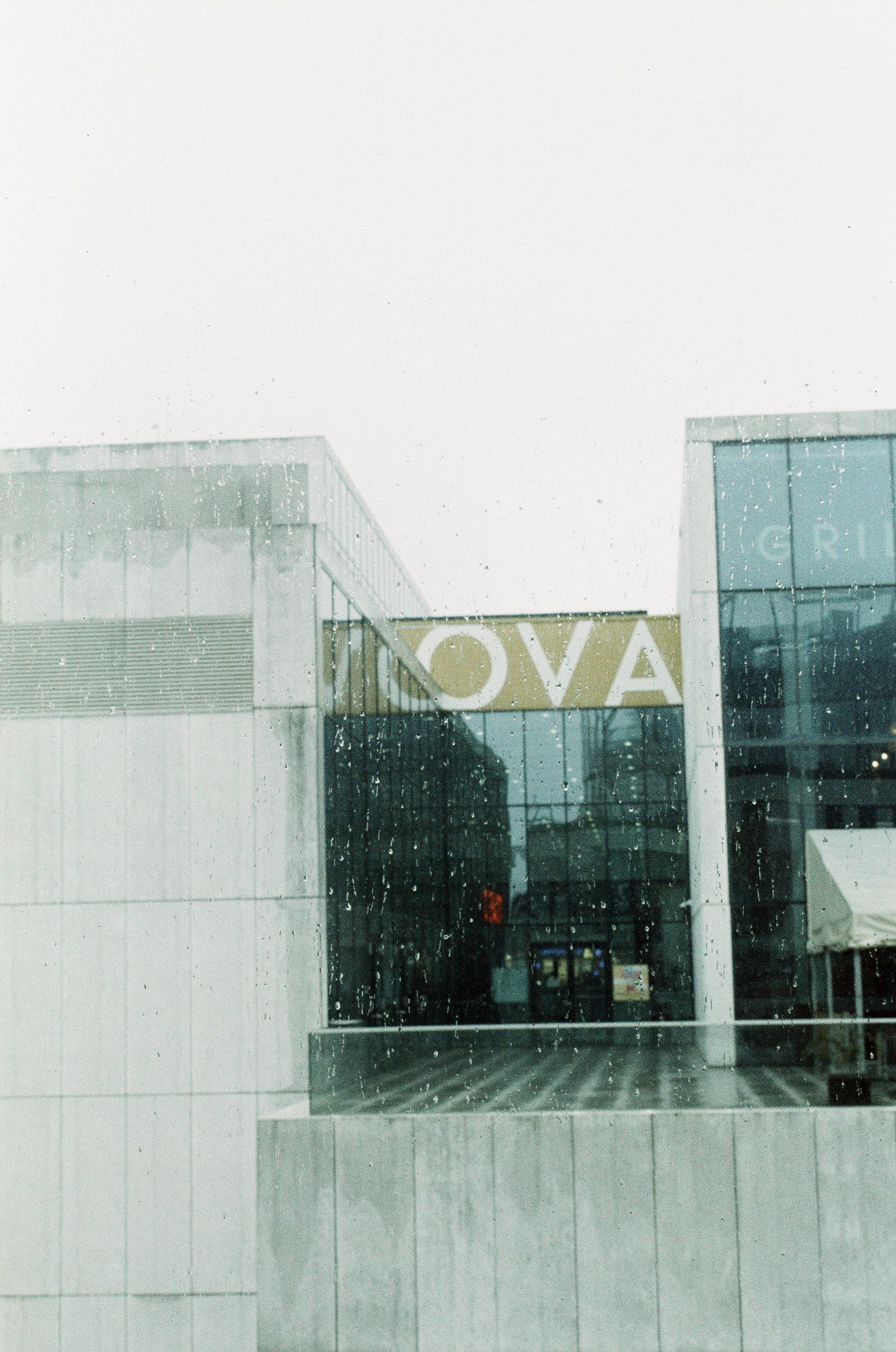 The image shows a modern building with glass and concrete elements. There is a sign with the letters "OVA" visible. It's raining, as evidenced by the raindrops on the glass in front of the camera. The architecture suggests it might be a commercial or office building.