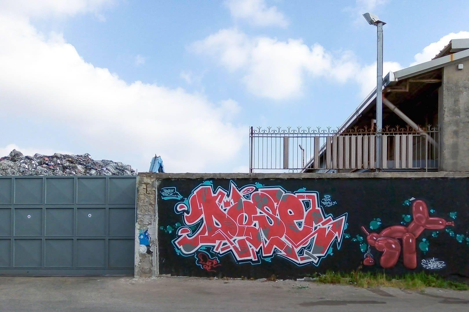 Graffito con lettere stilizzate rosse e l'immagine di un cane fatto di palloncini intento a fare i suoi disegni, realizzato sul muro di quella che sembra essere un impianto di trattamento dei rifiuti.