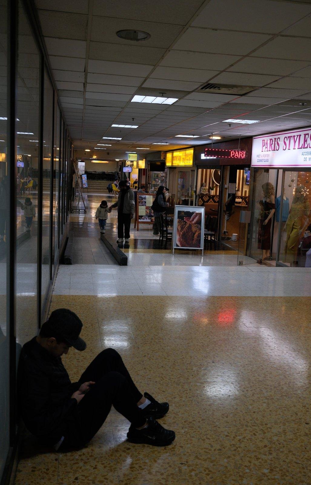 Une galerie marchande, avec au 1er plan, un jeune homme assis par terre, adossé à une vitrine, casquette sur la tête, en train de consulter son portable