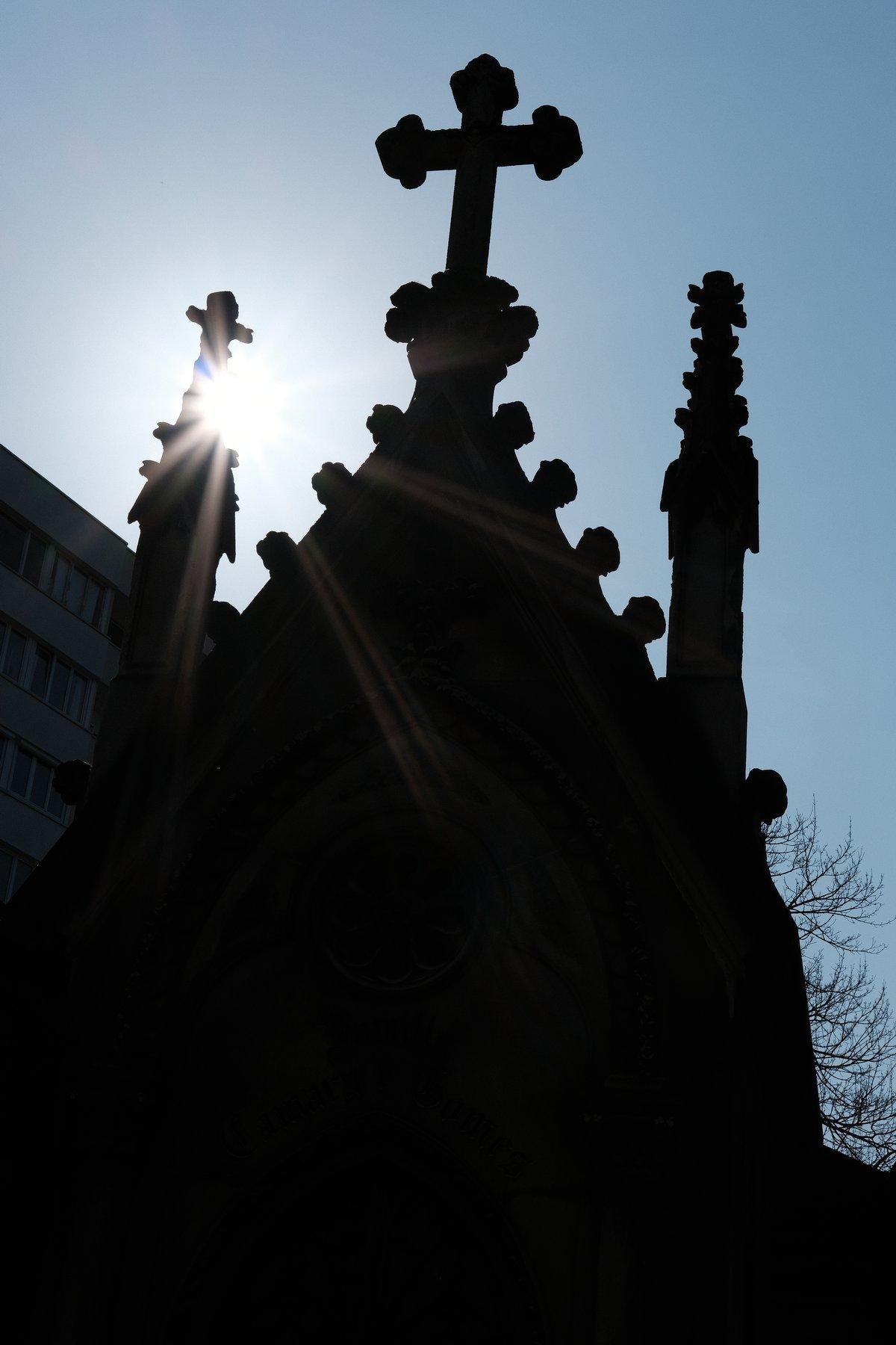 Photo verticale. Un mausolée et sa croix au sommet et deux autres plus petites sur les côtés, en contre-jour, le soleil perçant à côté de la croix de gauche, sur ciel bleu