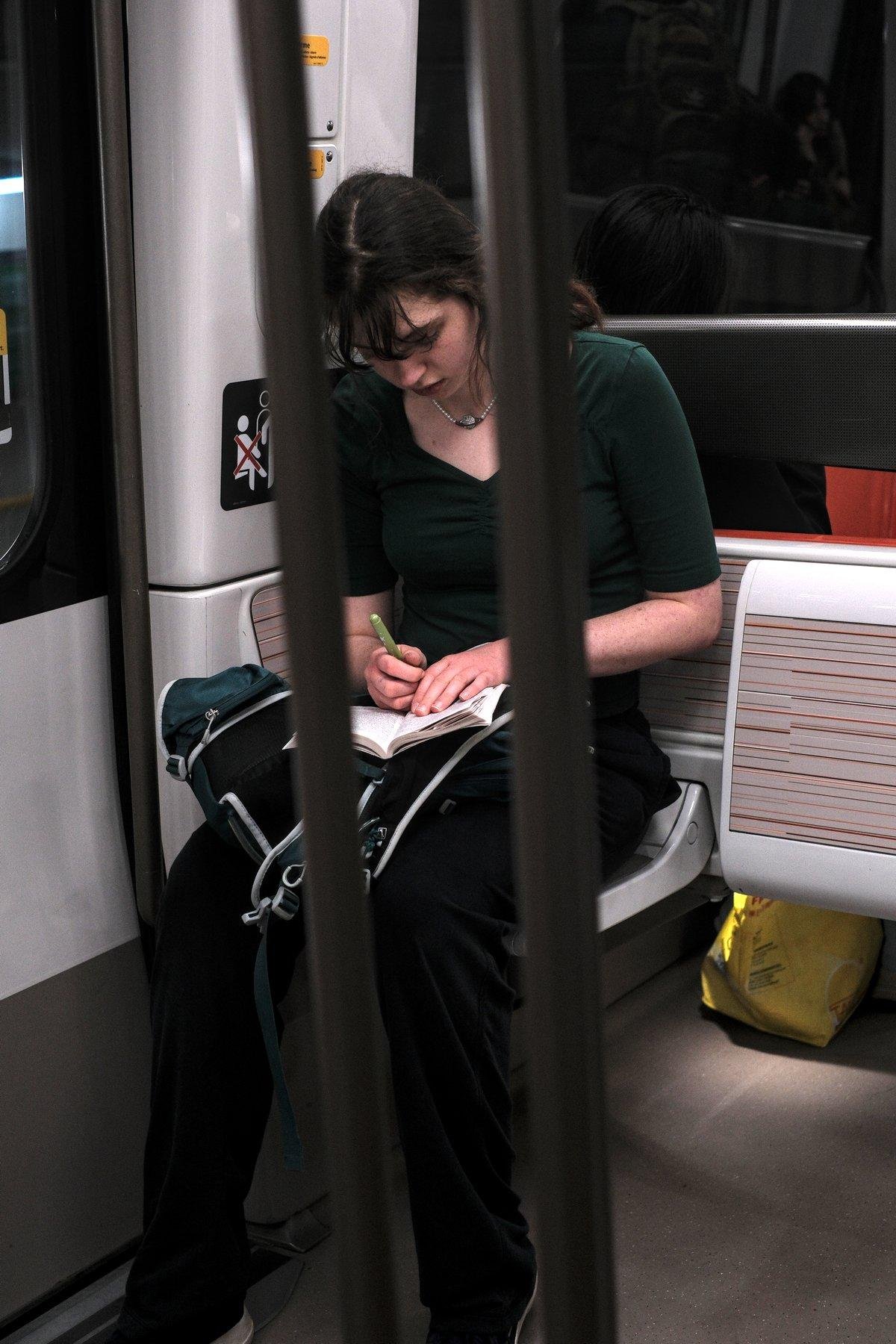 Photo verticale. Vue entre 2 barres de métro (celles faisant partie d’un trio au centre), une jeune femme brune aux cheveux longs attaché, collier, haut vert sombre, pantalon noir, écrit sur un carnet de notes posés sur un sac vert assorti au haut