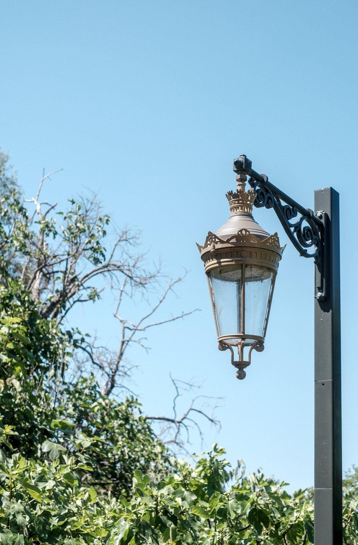 Photo verticale. Sur un fond de ciel bleu, un lampadaire et son attache ouvragé, la lampe sur des tons dorés et l’attache gris ardoise, et un poteau de la même couleur, mais de forme carré tout simple sans aucune fioriture