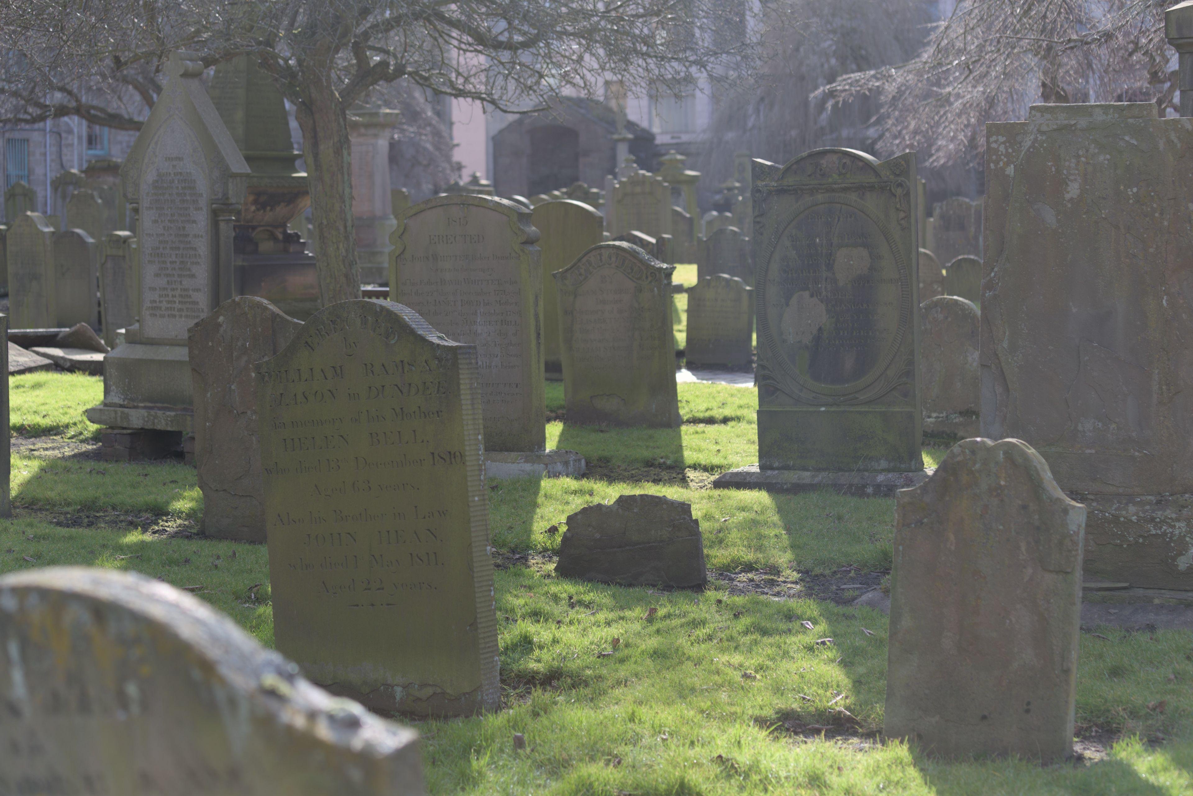 Photo format paysage de vieilles pierres tombales sans tombe apparente devant. Elles sont couvertes de mousse, la pelouse est épaisse et bien verte, un rayon de soleil les éclaire par derrière