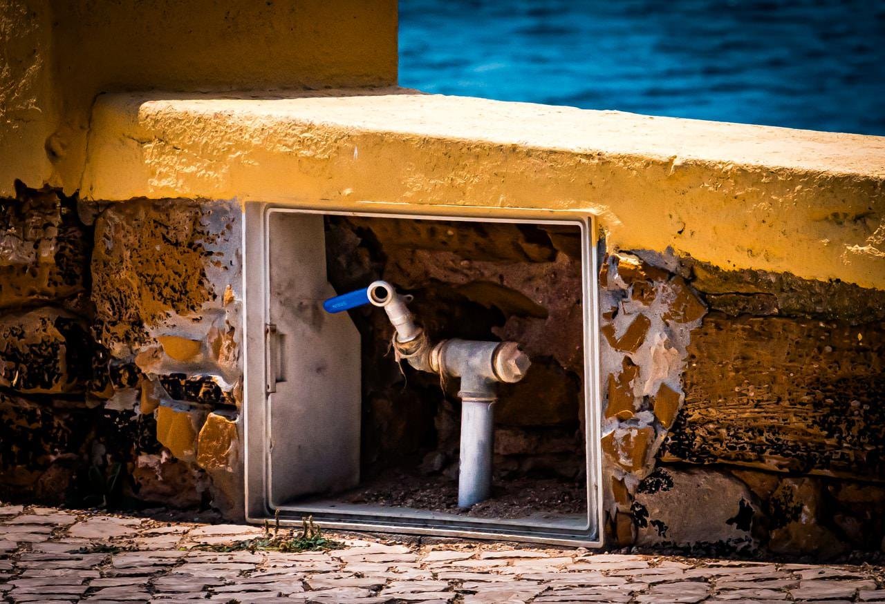 Yellow stone structure with a recessed metal compartment containing a water tap, featuring a blue handle. The compartment is set within a weathered wall situated near a body of water. Stone pavement is visible at the base.