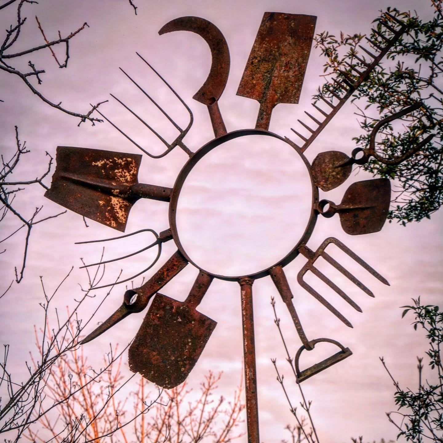 The image depicts a sculpture made from various rusted garden tools arranged radially around a circular frame, resembling a sun or a star. These tools include items like shovels, rakes, and pitchforks, and the sculpture is set against a sky background, framed by some tree branches.