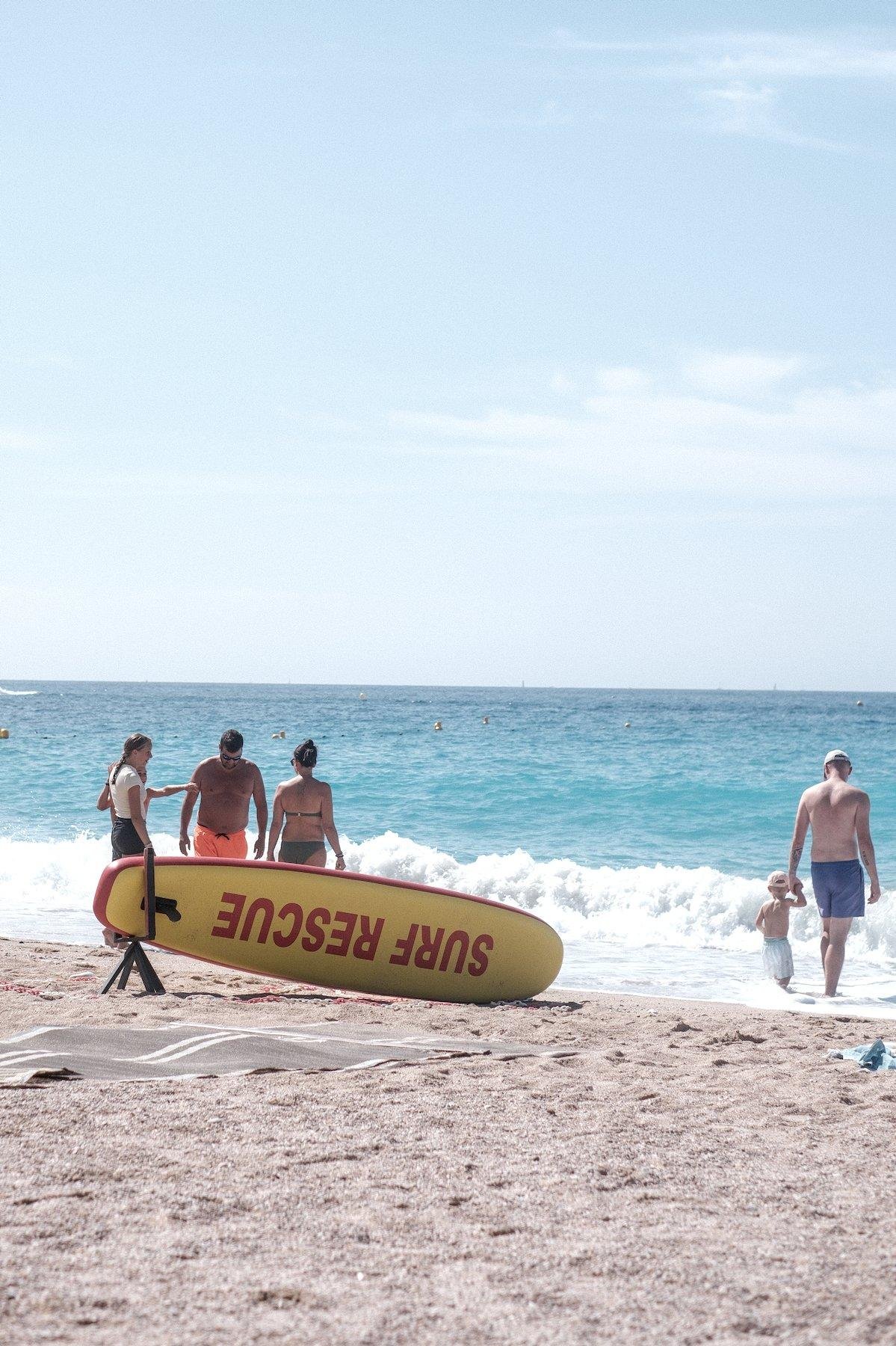 Photo verticale. Sur un plage de sable, près de l’eau, une planche de surf jaune et rouge marquée en rouge avec les mots « Surf Rescue » est posée sur la tranche sur un petit trépied noir. Autour, des gens trempent leur pieds dans l’eau