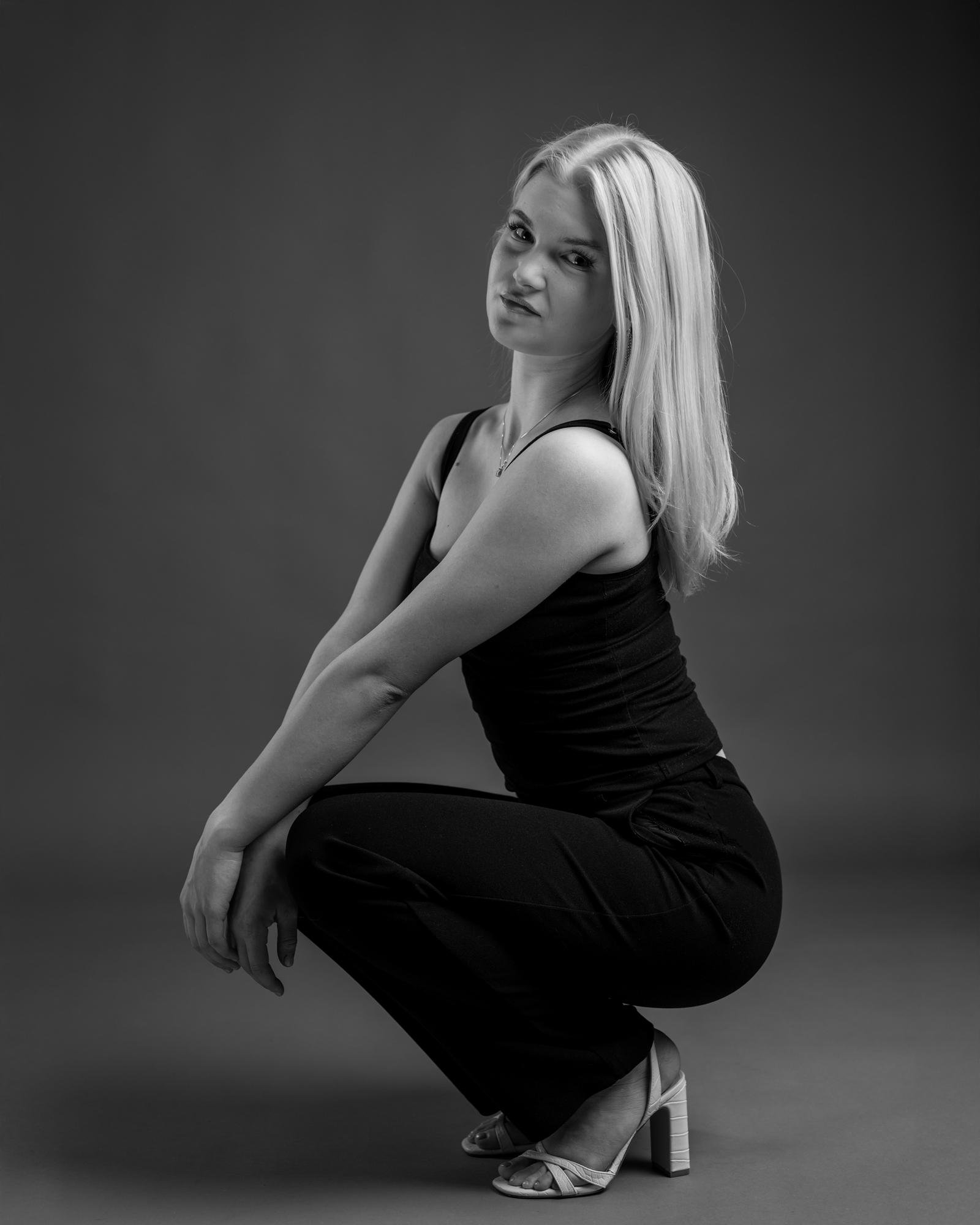 This is a black and white photograph of a person with long hair, wearing a sleeveless top, pants, and high-heeled shoes. The person is squat or crouch posing, looking towards the camera. The background is plain and the focus is on the person in the image.
