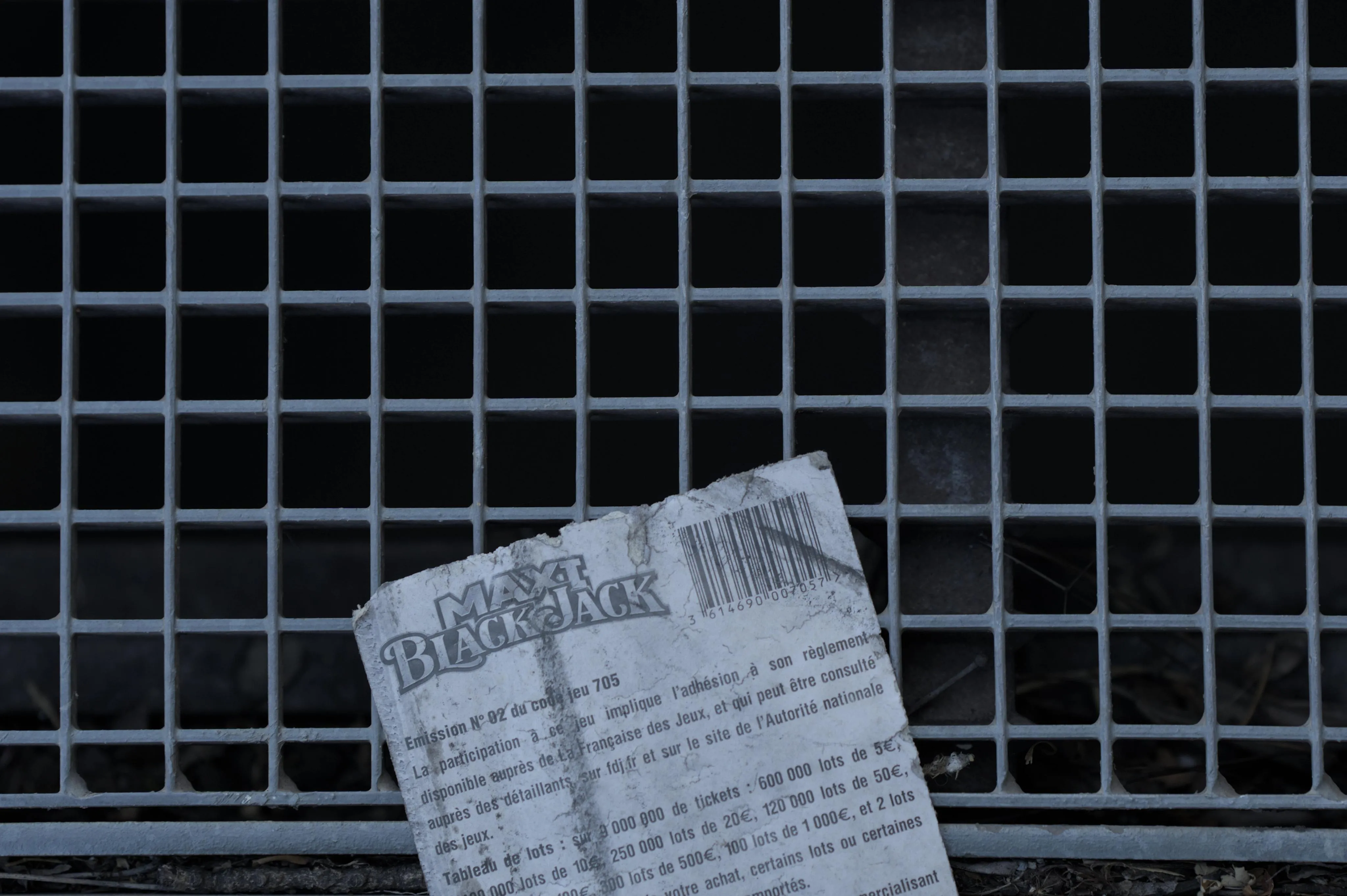 Close-up photograph on a metal grate with a dirty, withered paper ticket on it. The paper ticket is titled "Maxi blackjack" and includes a barcode as well as written rules and requirements for gambling.