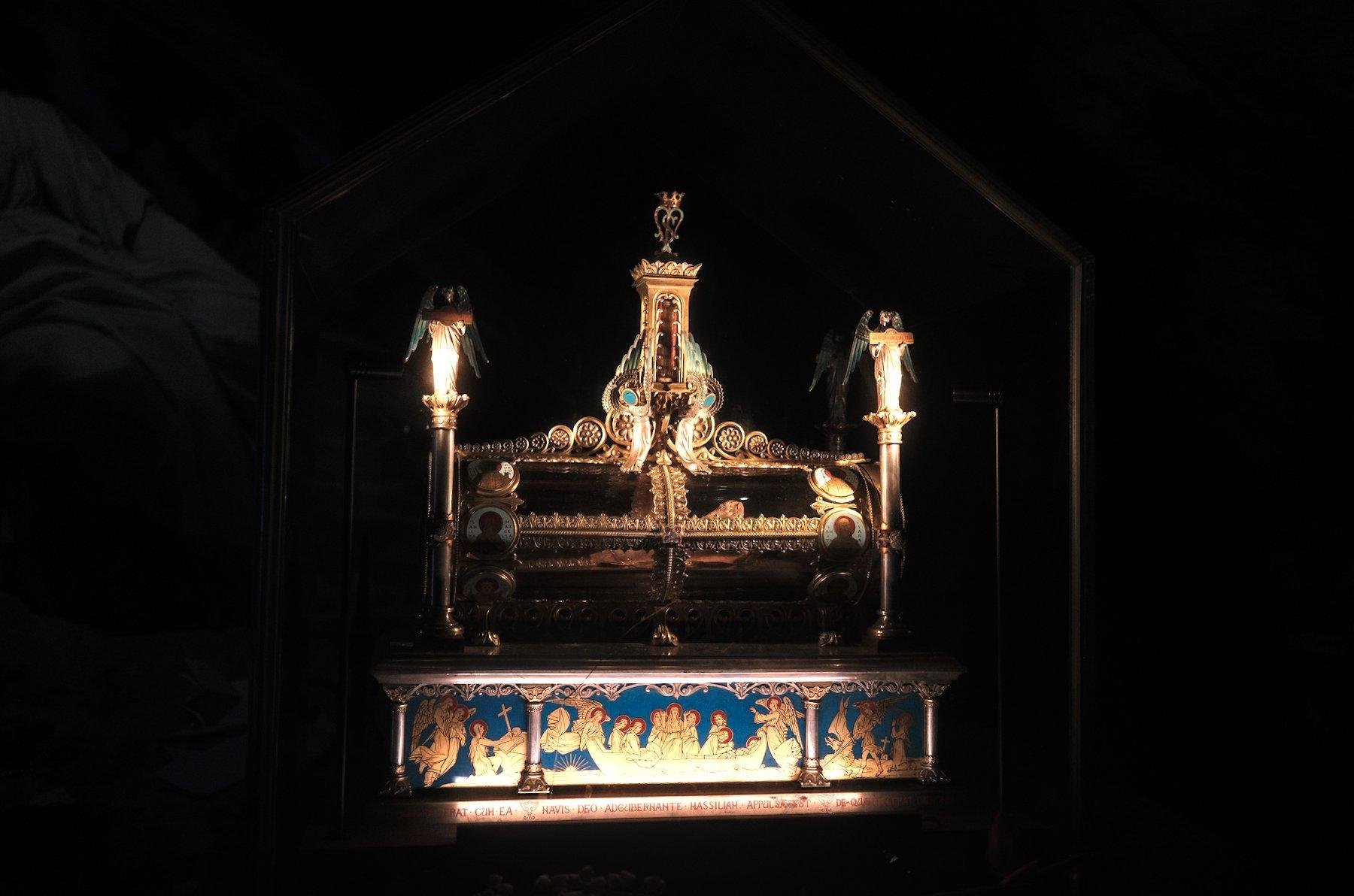 Photo horizontale. Dans un lieu très sombre, le luxueux reliquaire du fémur de Marie-Madelaine, tout dorée avec des touches de bleu royal, éclairé par en dessous
