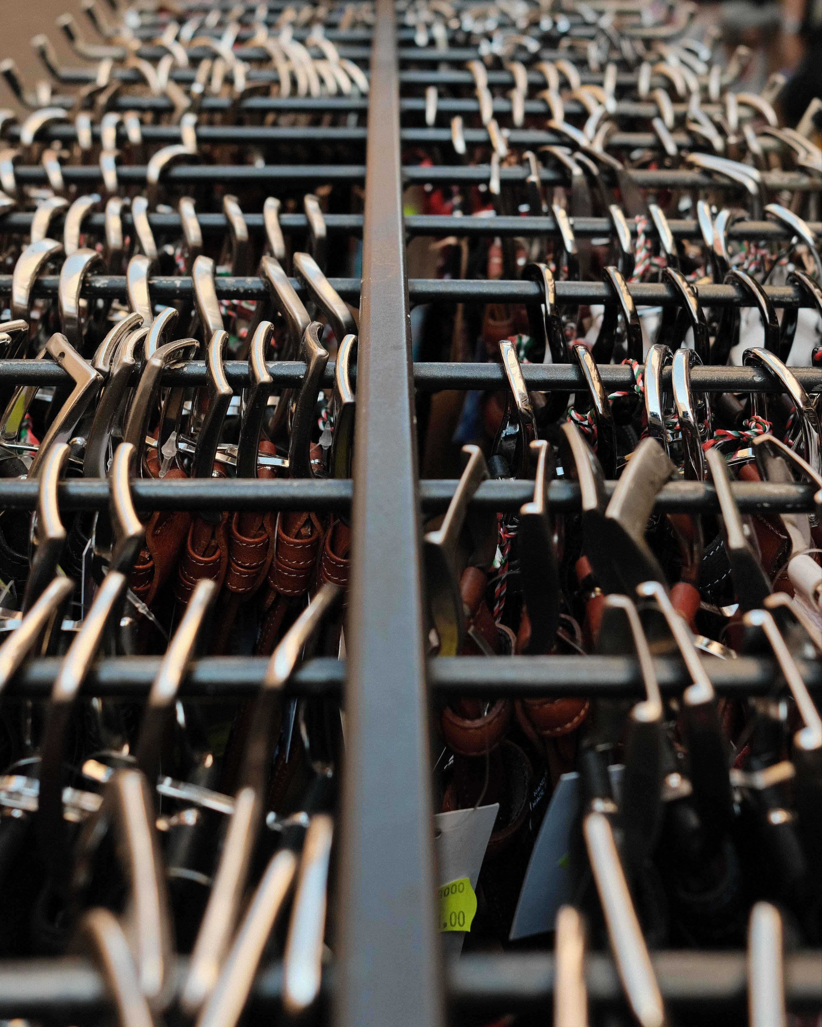 Rows of metal belt buckles packed tightly on black rails, separated by a central divider, photographed from an elevated perspective. The belts below are barely visible, instead the tightly paved metal buckles create an interesting pattern.