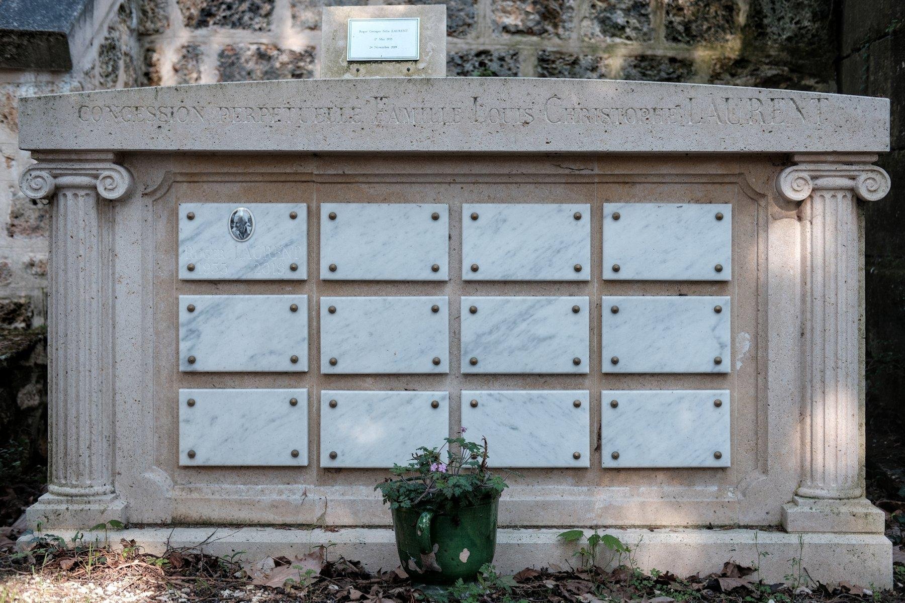 Photo horizontale. Un tombeau de face, en forme de temple grec, avec des colonnes de chaque côté. Sur la façade, une grille de plaque de marbre blanc de 4 colonnes et 3 lignes. Seule celle en haut à gauche est gravée et porte un petit portrait dans un médaillon.