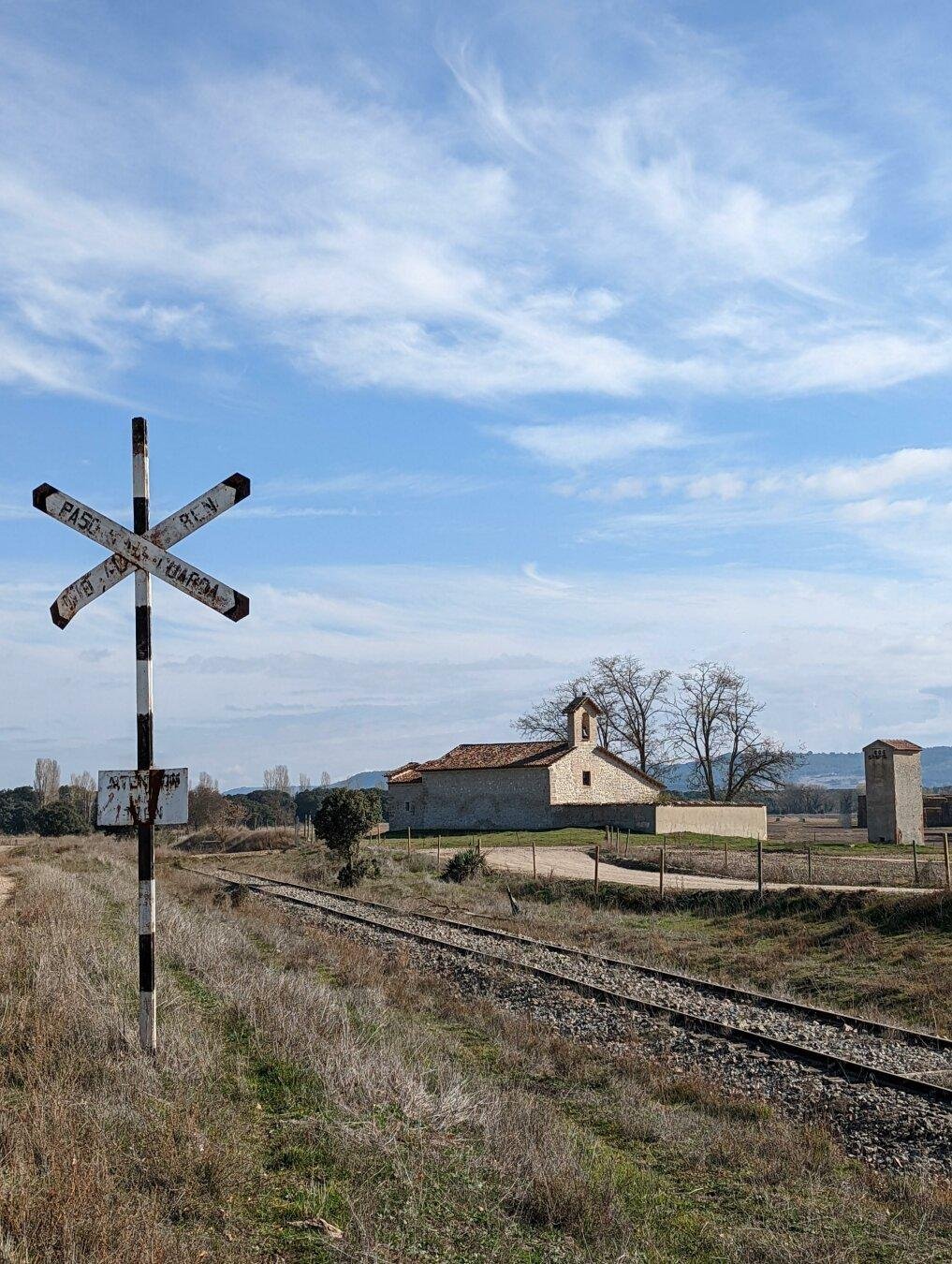 Paisaje otoñal. En primer plano, se ve hierba y una señal ferroviaria antigua de paso a nivel, en forma de equis en blanco y negro. En segundo plano, pasa una vía de tren antigua, que cruza la foto de derecha a izquierda hasta perderse. Al fono, lo que parece una pequeña iglesia o ermita. El cielo azul está salpicado por algunas nubes de aspecto sedoso. En suma, la foto transmite tranquilidad, con la señal como protagonista.