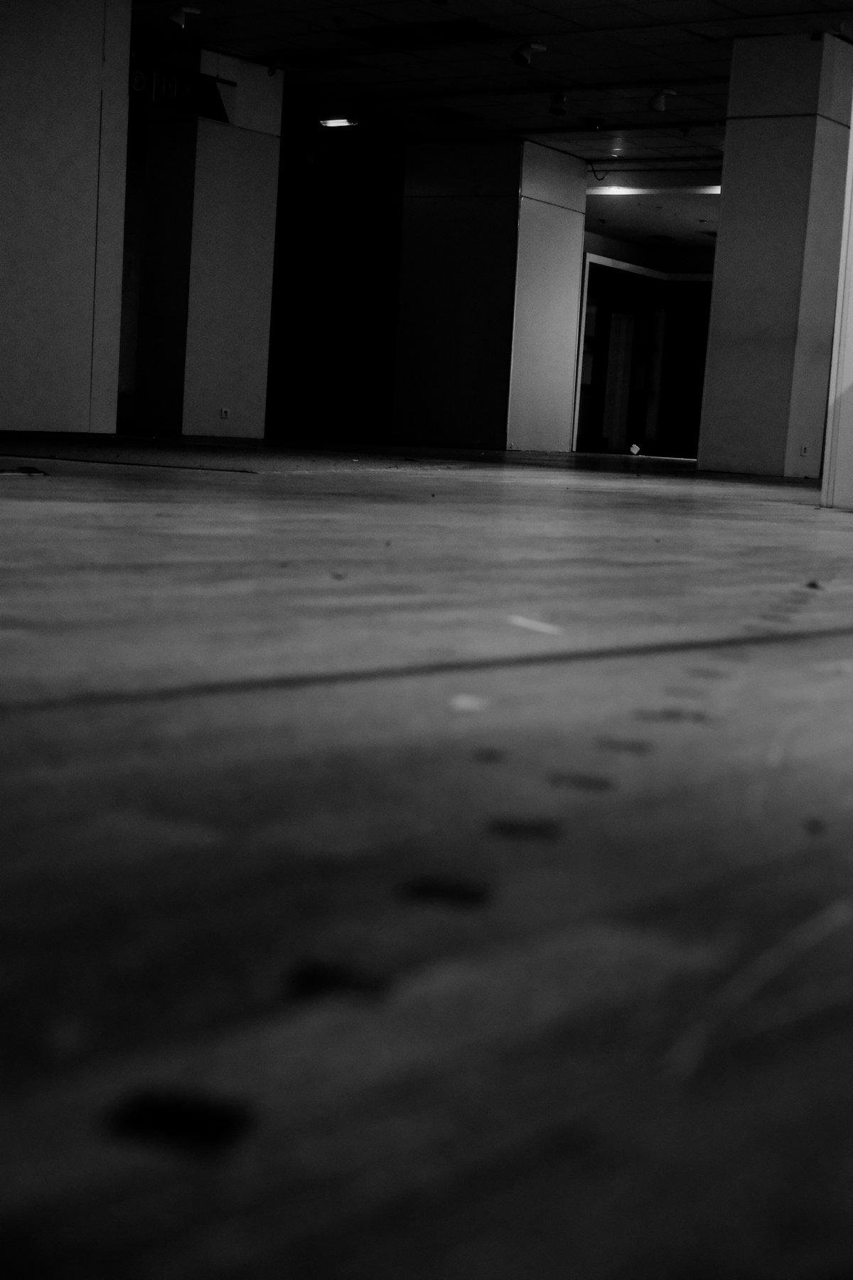 Photo verticale en noir et blanc. Vue à ras du sol en parquet qui a un marquage avec des petits carrés noirs partant du coin SW et croisant le bord Est de l’image à un peu au dessus de la moitié. On voit de faibles lampe au loin, un pilier carré, des murs vides et on remarque l’absence de mobilier
